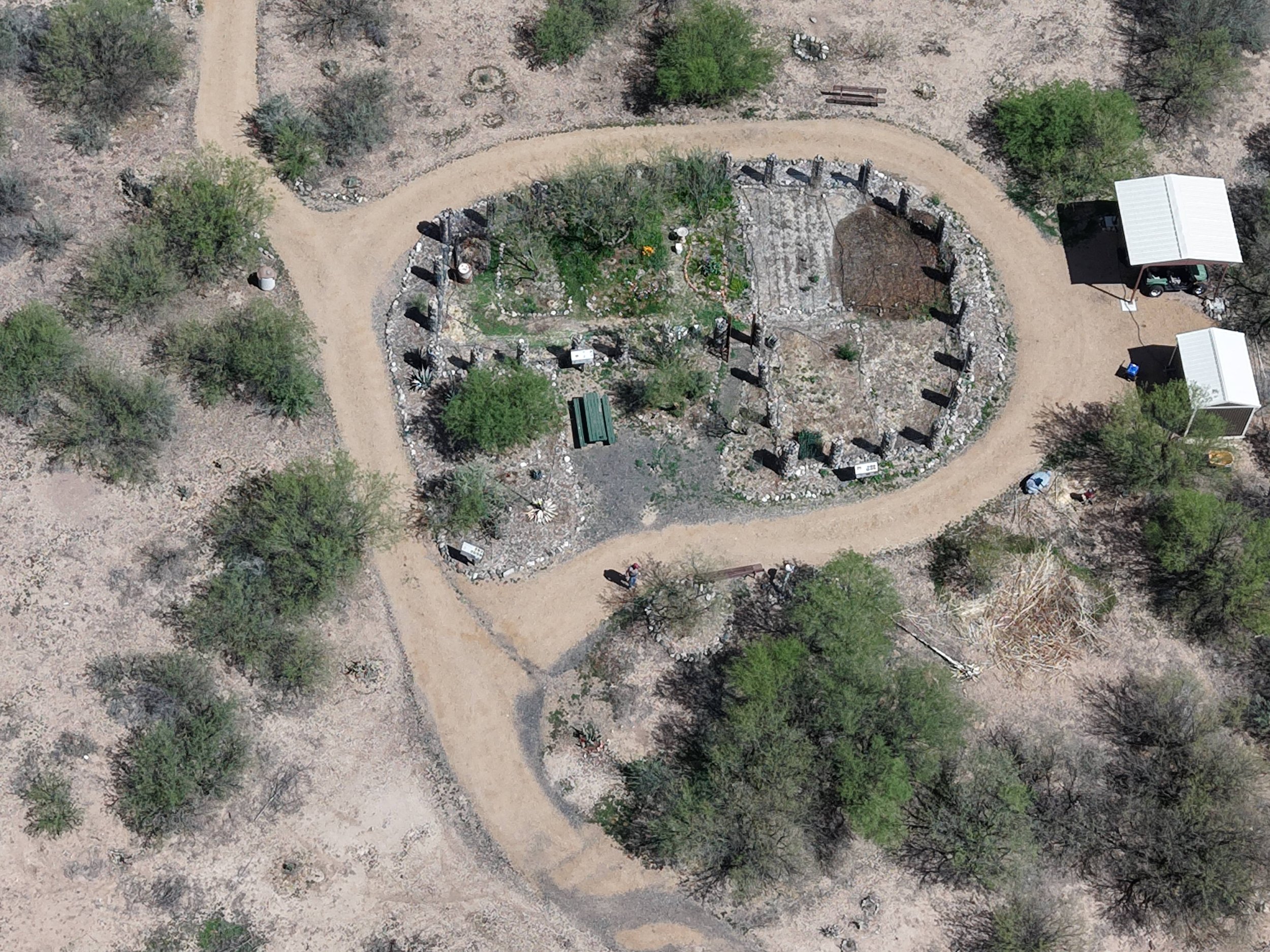Aerial view of a desert landscape with a circular garden surrounded by rocks and plants. There are pathways, trees, and two small buildings nearby.