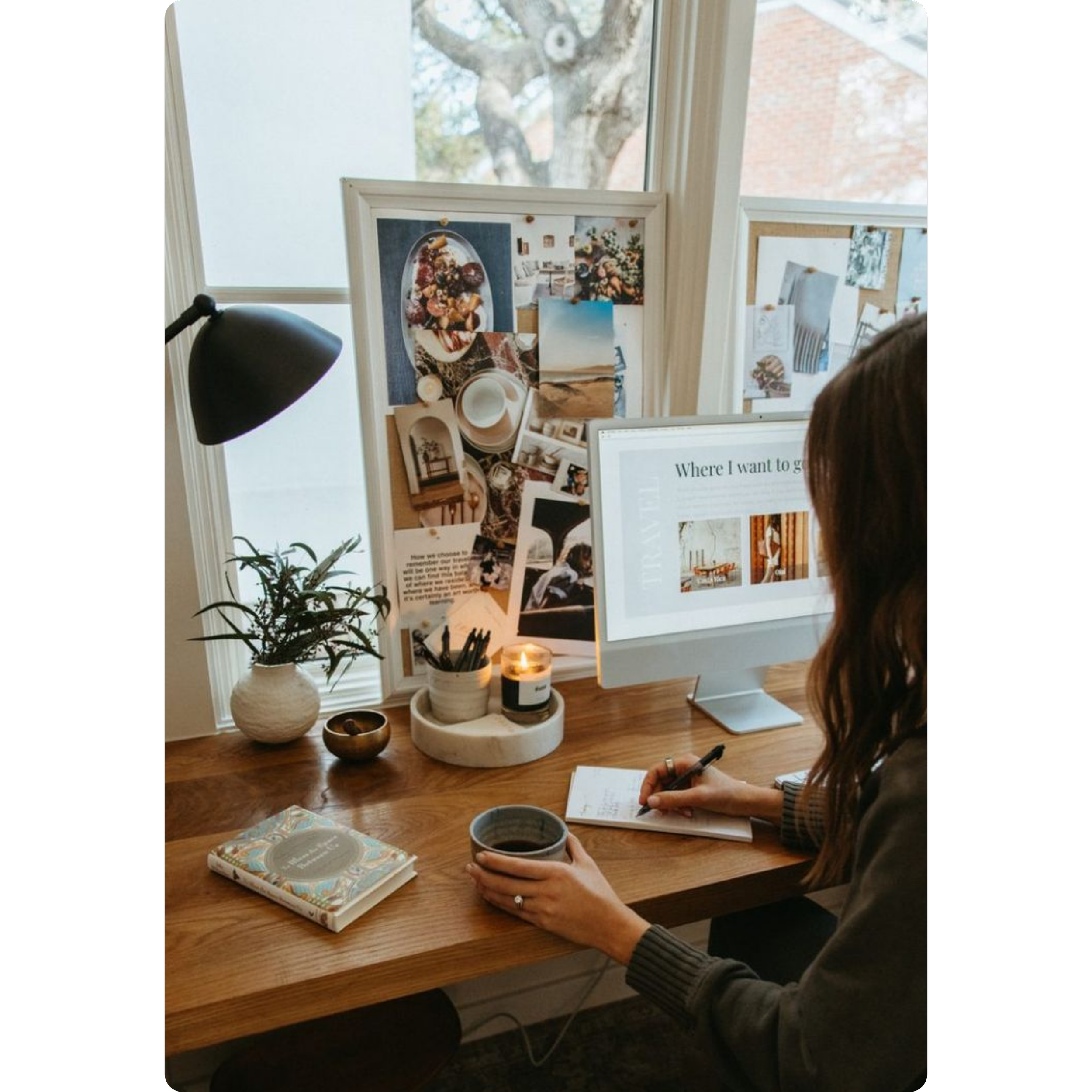A woman seated at a wooden desk working on a computer with a large monitor that displays text and images. The desk has a potted plant, a candle, notebooks, and writing utensils. Behind her, there are two whiteboards covered with photos, illustrations, and notes. A window with a view of a large tree is visible in the background.