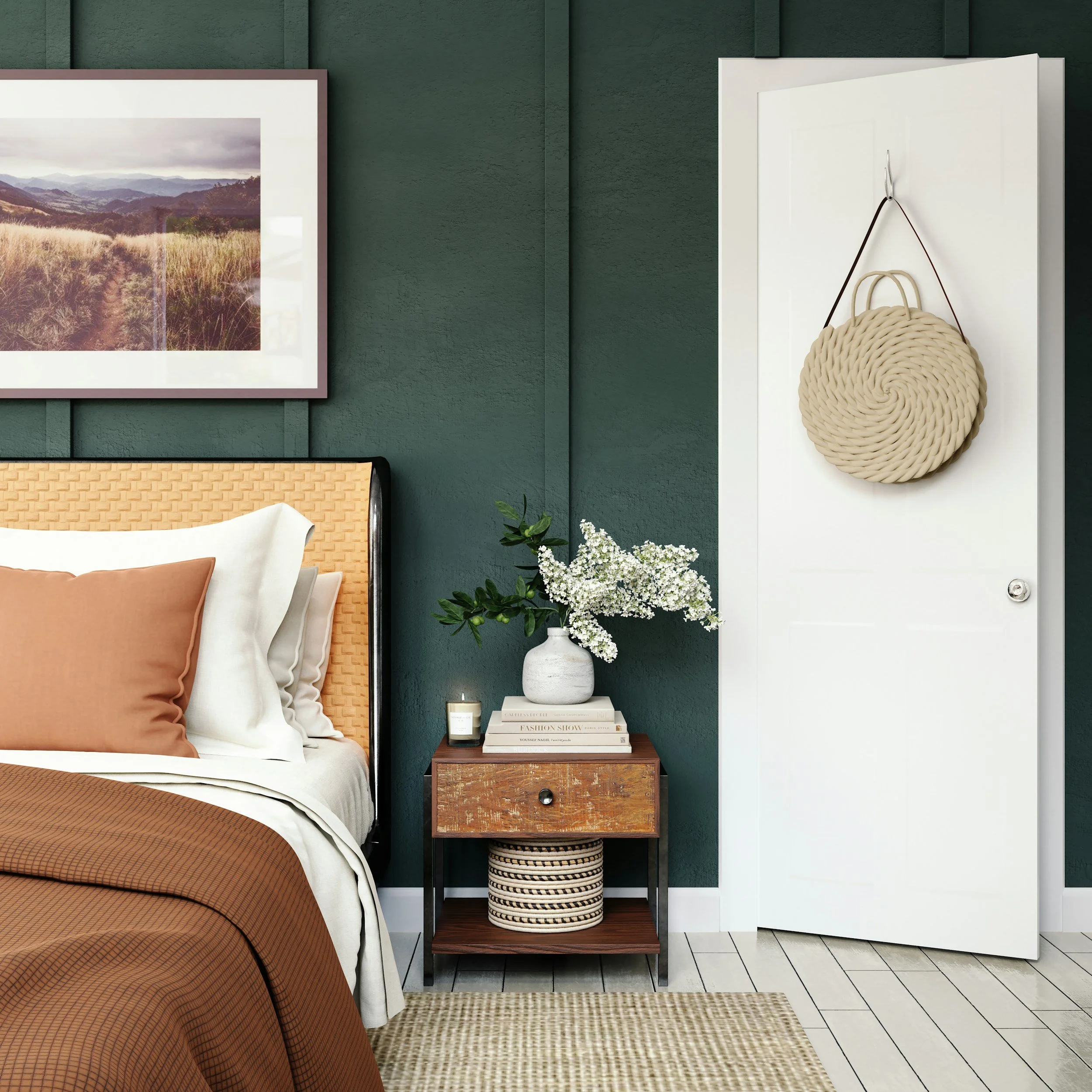 A bedroom corner with a bed featuring an orange-brown headboard and pillows, a wooden nightstand holding a white vase with white flowers and a small candle, green textured wall, framed landscape photo on wall, and white door with a woven beige round bag hanging on it.