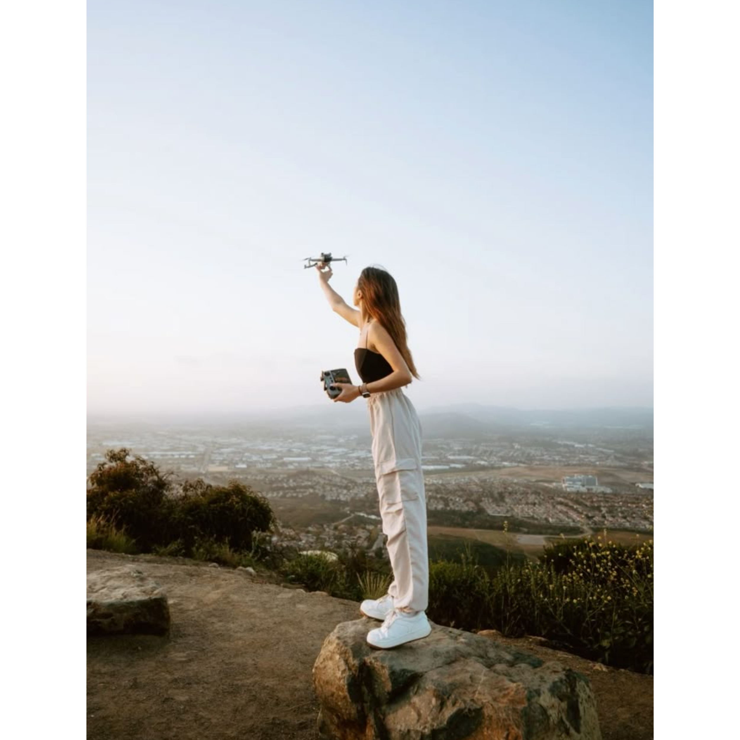 A woman standing on a rock at a scenic overlook, flying a drone with a remote controller in her other hand, against a backdrop of a vast landscape and open sky.