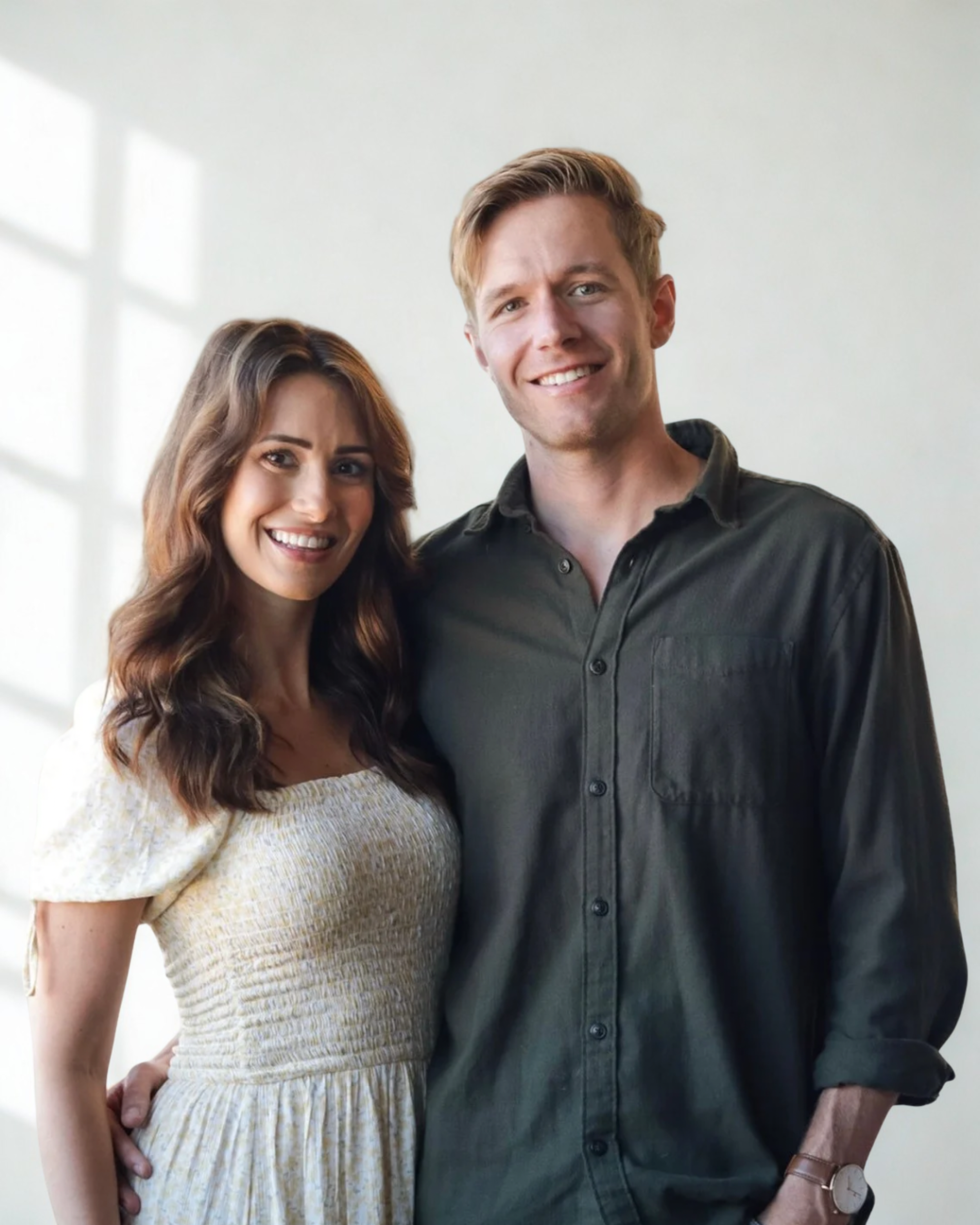 A smiling couple standing close together indoors, with sunlight coming through a window behind them.