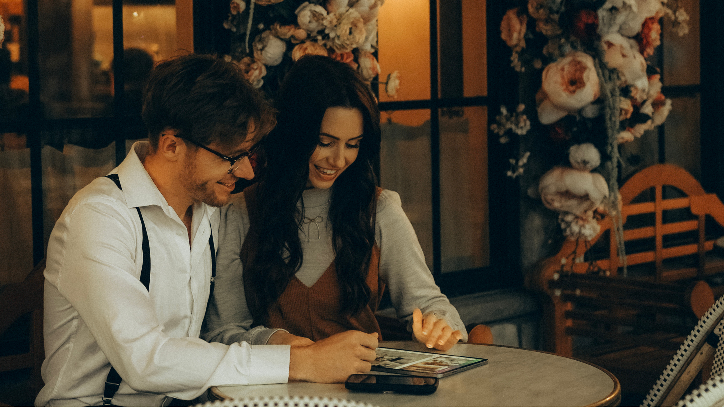 A man and woman sitting at a table in a warm, cozy restaurant, looking at a tablet and smiling together.