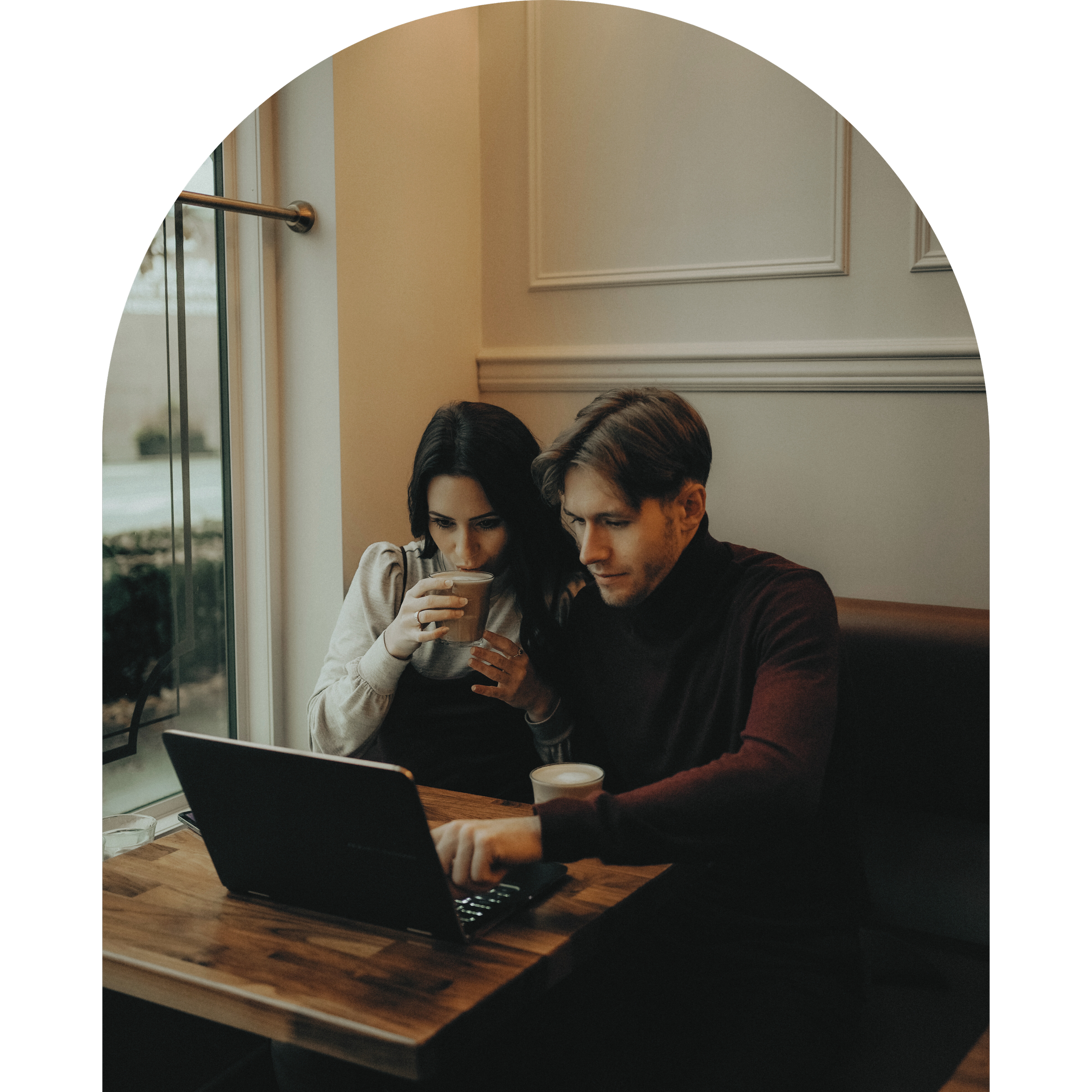 A man and woman sitting at a wooden table in a cafe, looking at a laptop, with the woman drinking coffee.