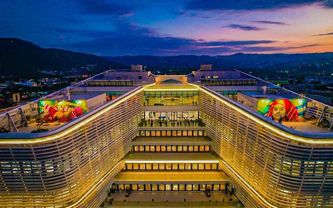 BINAES, El Salvador, library, city of San Salvador Aerial view of a modern multi-story building at dusk with illuminated edges, featuring murals and windows, surrounded by a cityscape and mountains in the background.