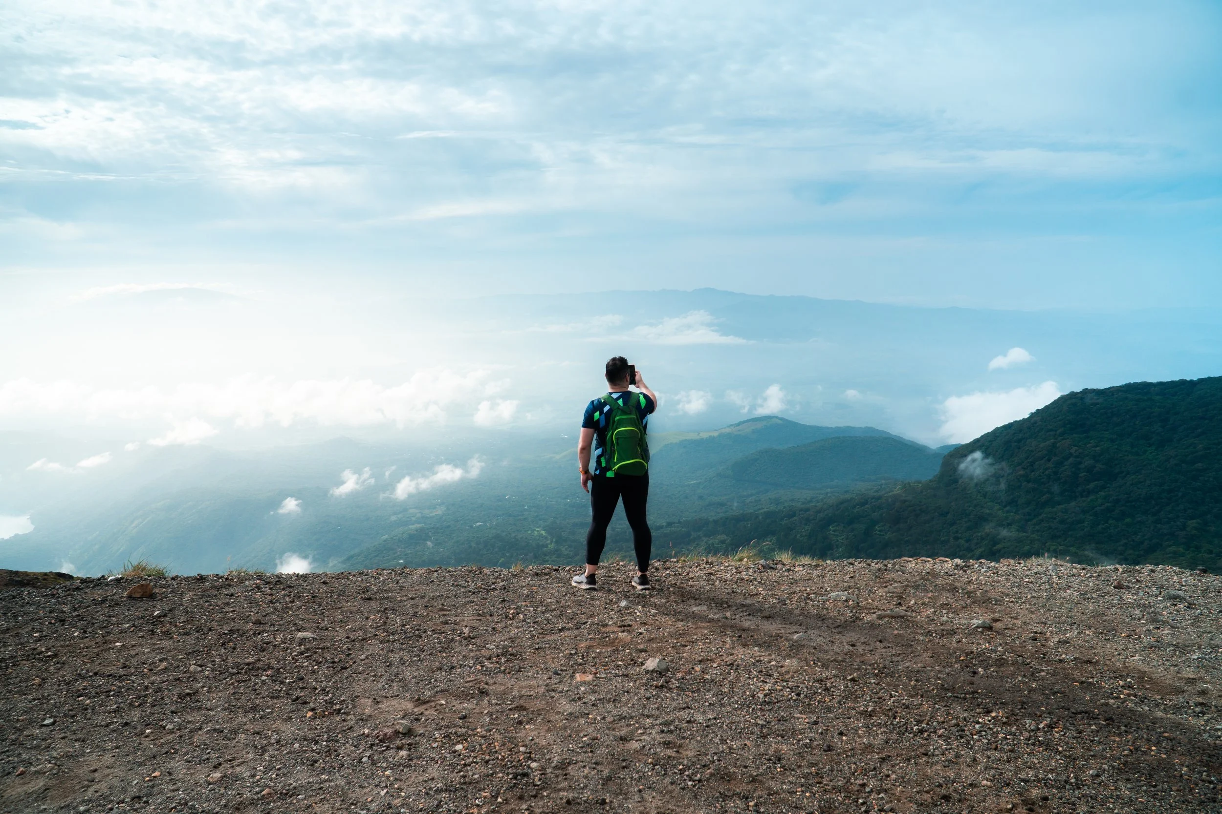 A person standing on a mountain trail, facing away, looking at a view of green mountains and clouds with a blue sky.