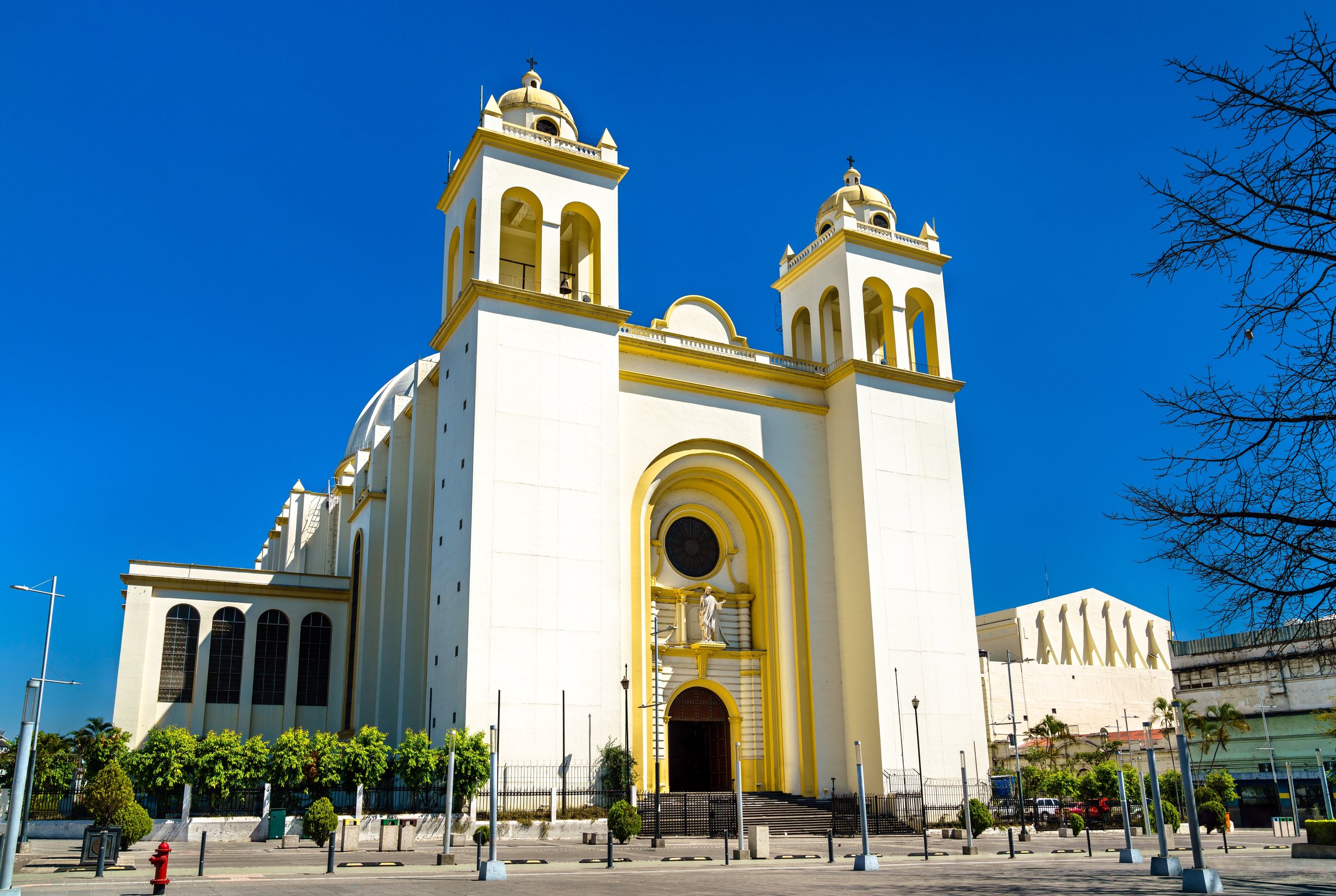 A large white church with two towers with crosses on top, set against a clear blue sky. The church has a grand entrance with an arched doorway and a statue above it. There are small trees and street lamps in the foreground.