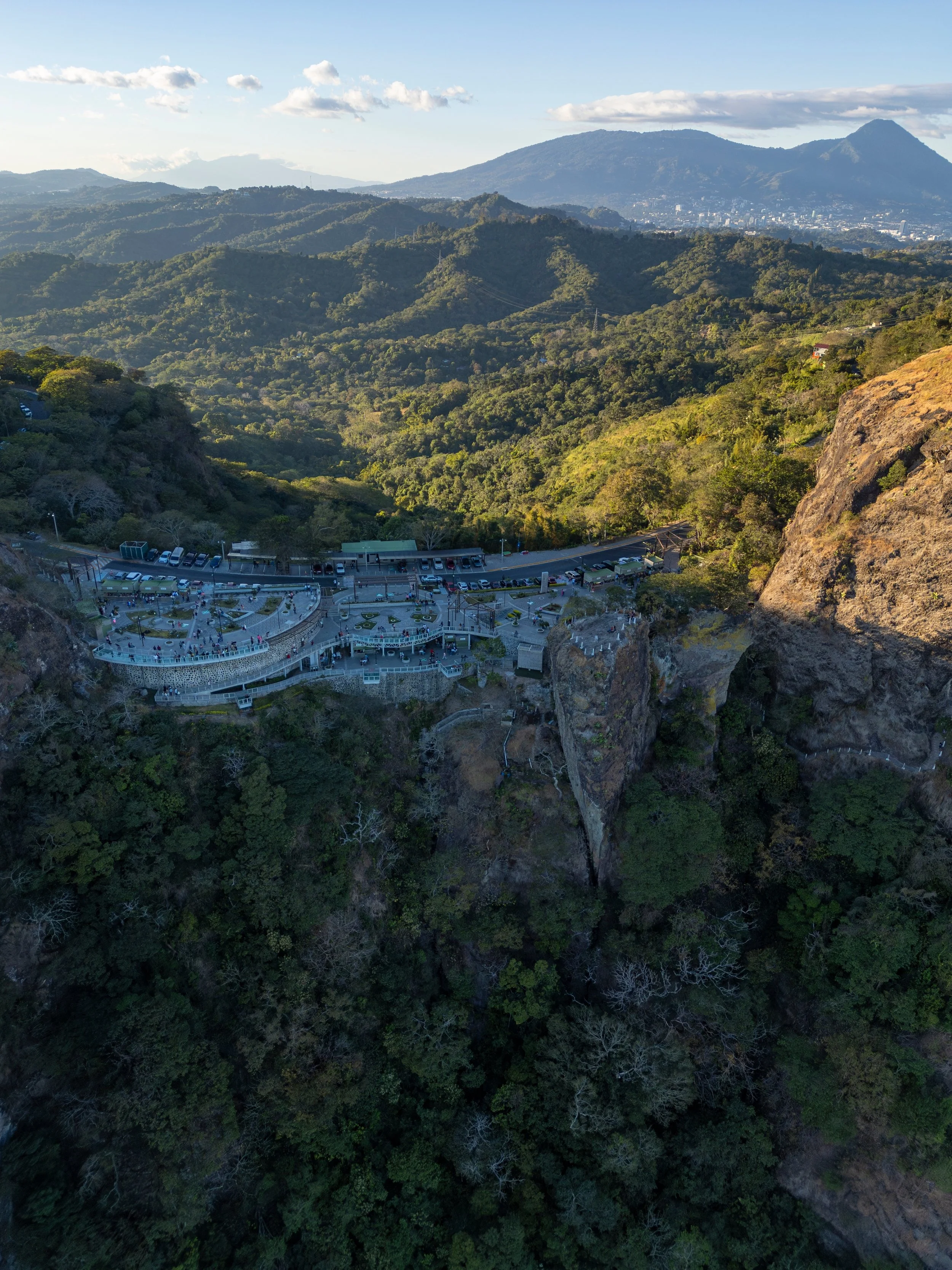 Aerial view of a mountainous landscape with lush green forests and a rocky cliff, a parking lot, and a viewing platform in the foreground, with mountains and a cityscape in the background.