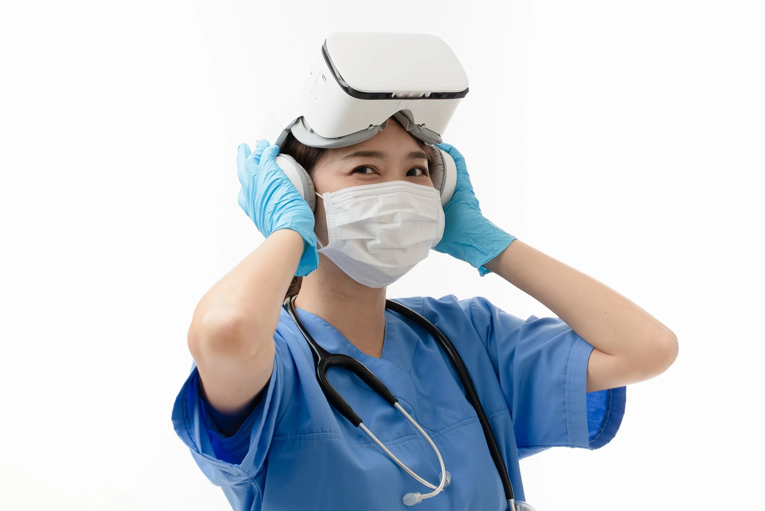 A healthcare worker wearing a blue scrub top, mask, gloves, and stethoscope placing a virtual reality headset on her head.