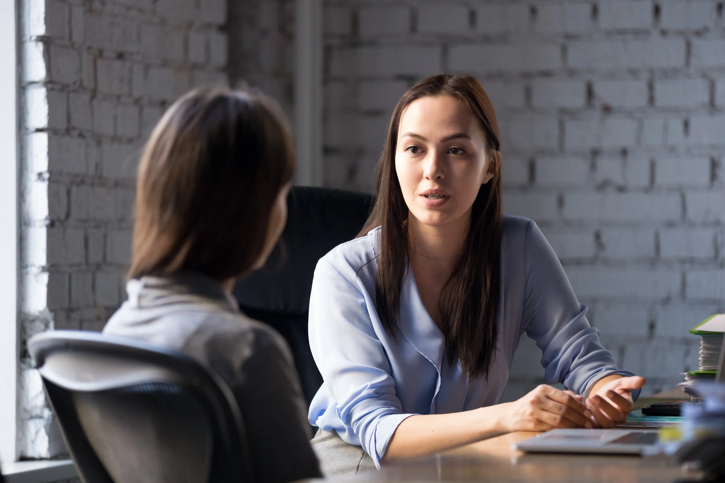 Two women having a conversation in an office setting with a white brick wall background.