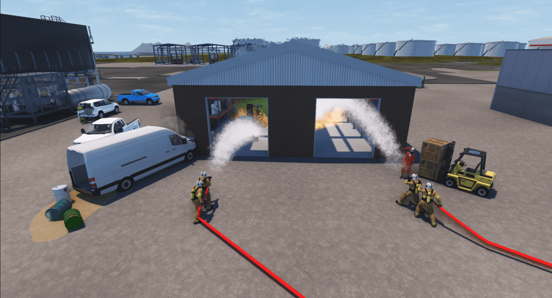 Firefighters at a training facility practicing fire suppression, with water hoses spraying water into burning structures inside a large open warehouse, surrounded by emergency vehicles and equipment.