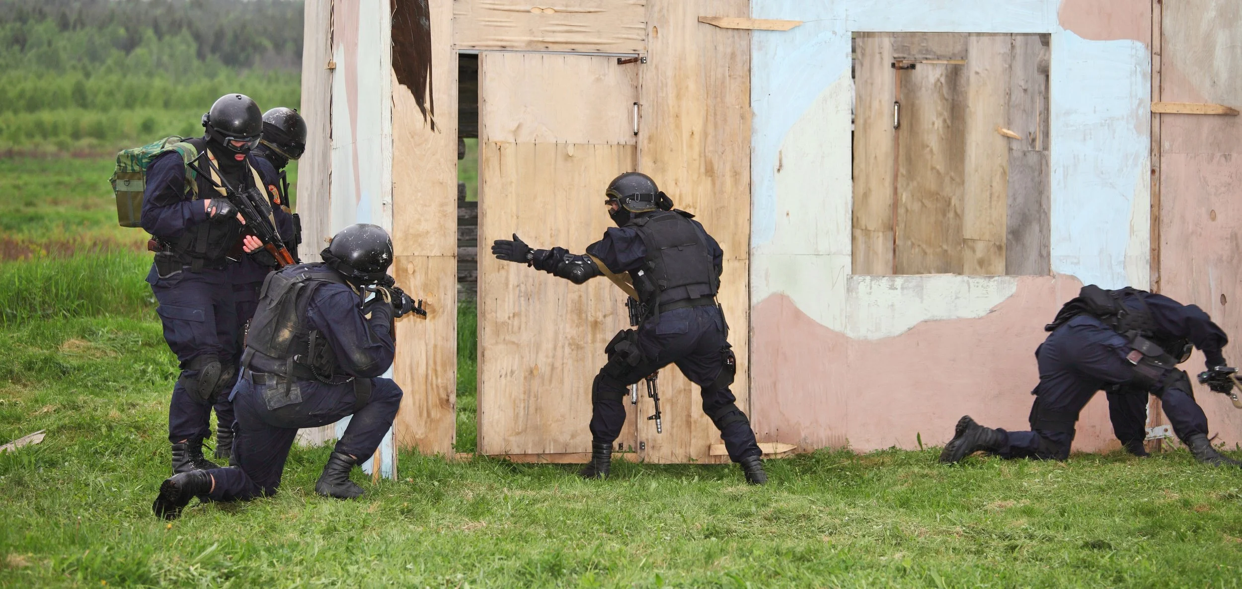 Five tactical police officers in black uniforms and helmets conducting a training exercise with a wooden structure and open door, some kneeling, one pushing against the wall, on a grassy field.