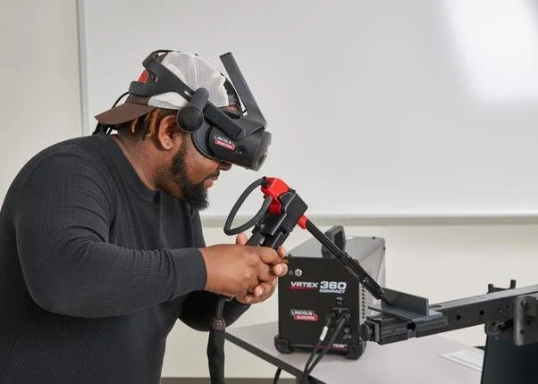 Person using a virtual reality headset and a welding tool in a classroom setting.
