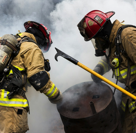 Two firefighters in full gear working together to extinguish a fire with smoke and ash around them.