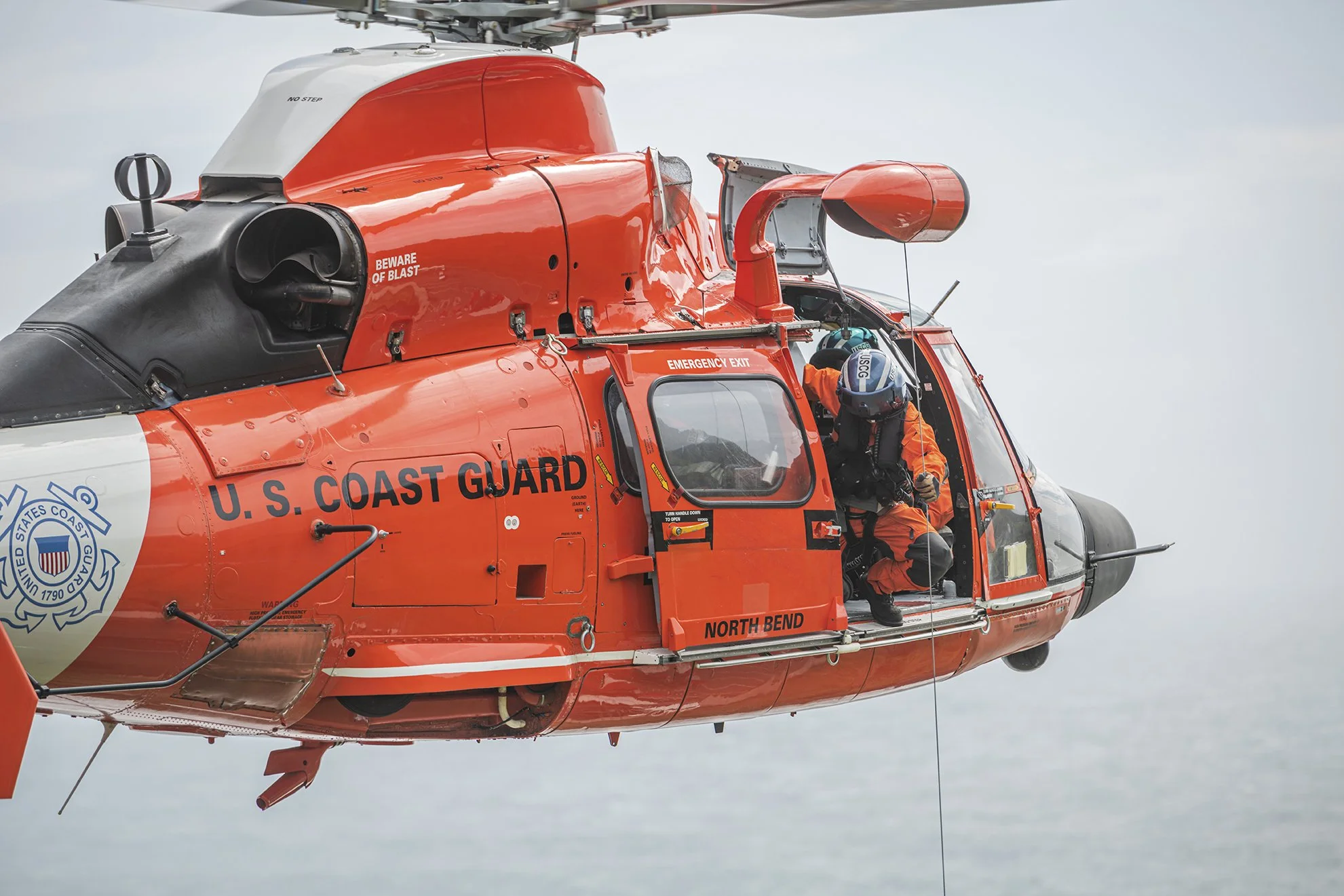 A U.S. Coast Guard helicopter flying in the sky with a crew member preparing for a rescue operation.