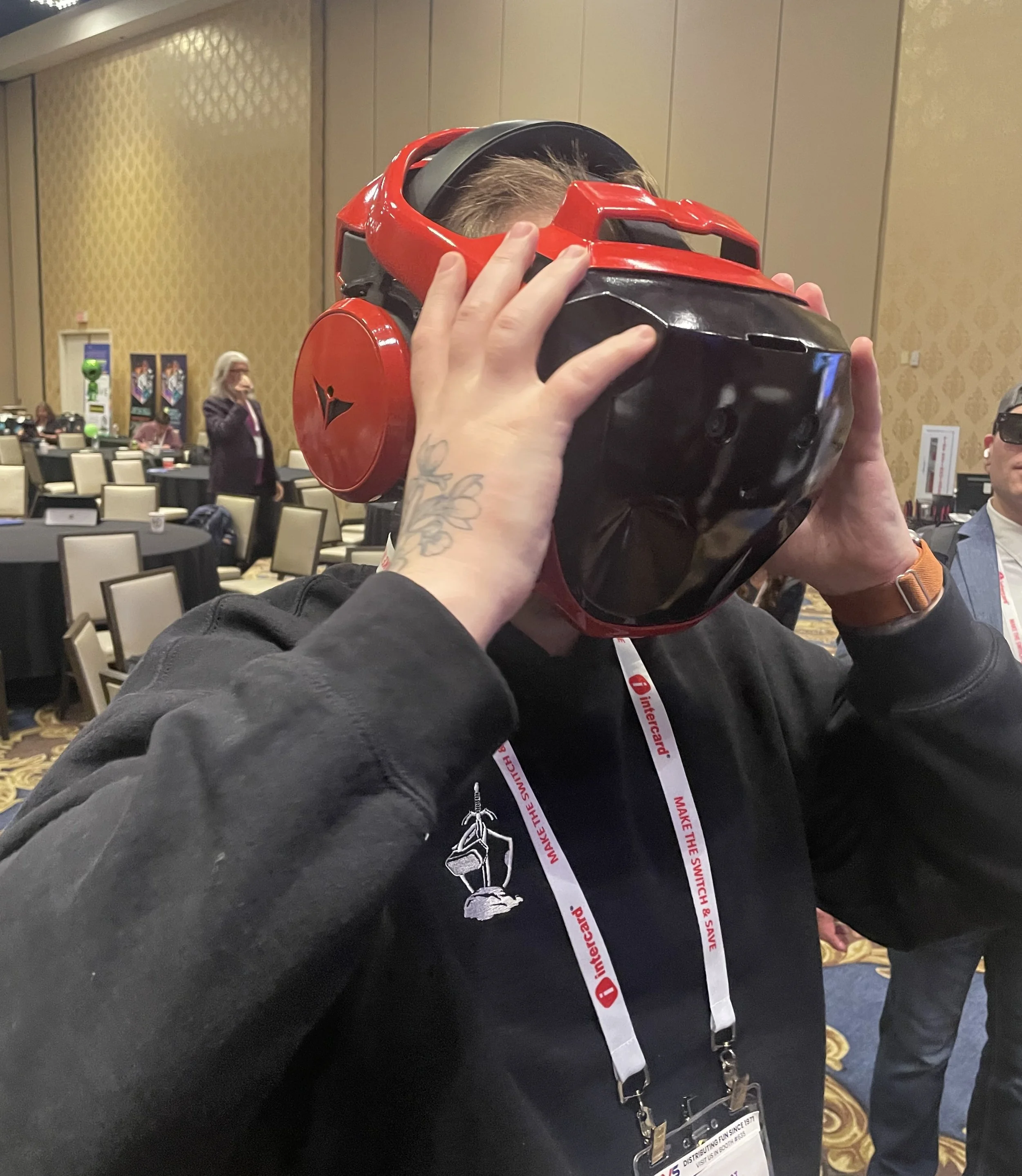 A person is trying on a large, black and red virtual reality helmet at a conference, with other people and tables in the background.