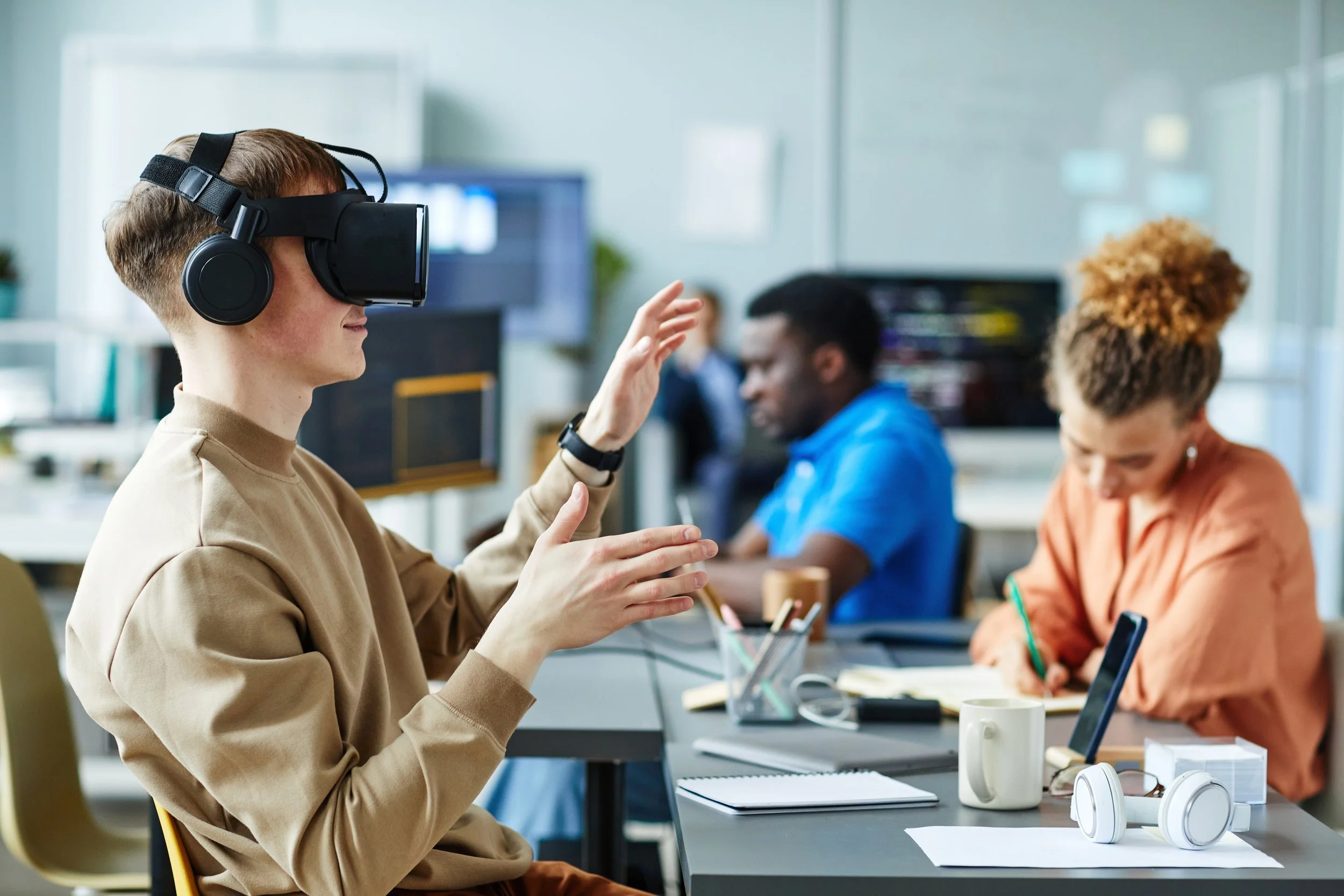 A young man wearing a virtual reality headset and headphones engages in a virtual experience in a modern office. Two colleagues work at their desks nearby, with office supplies, mugs, and a laptop visible on the tables.