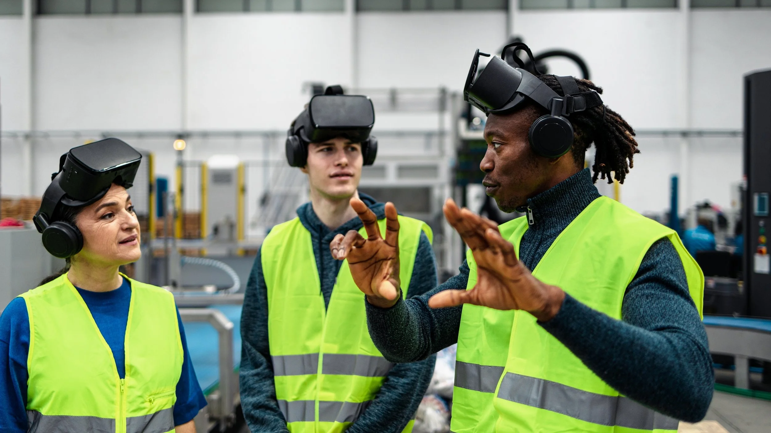 Three factory workers wearing virtual reality headsets and high-visibility safety vests in an industrial workspace, with one person explaining something while the others listen.