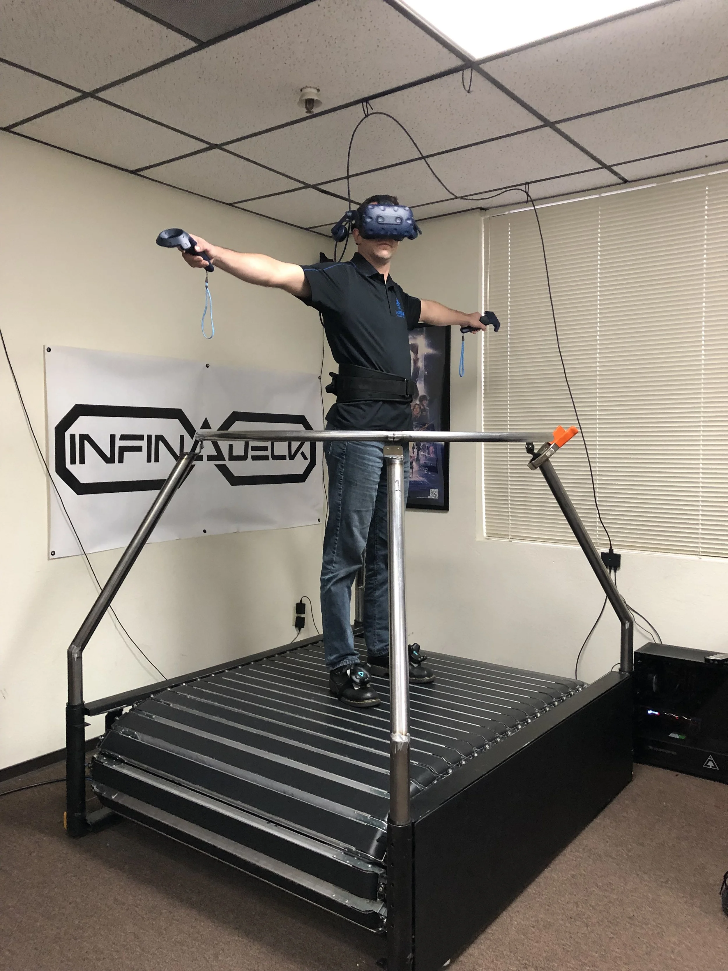 Man using virtual reality equipment while standing on a motion platform, with arms extended and wearing a VR headset, in an indoor room with a banner and blinds.