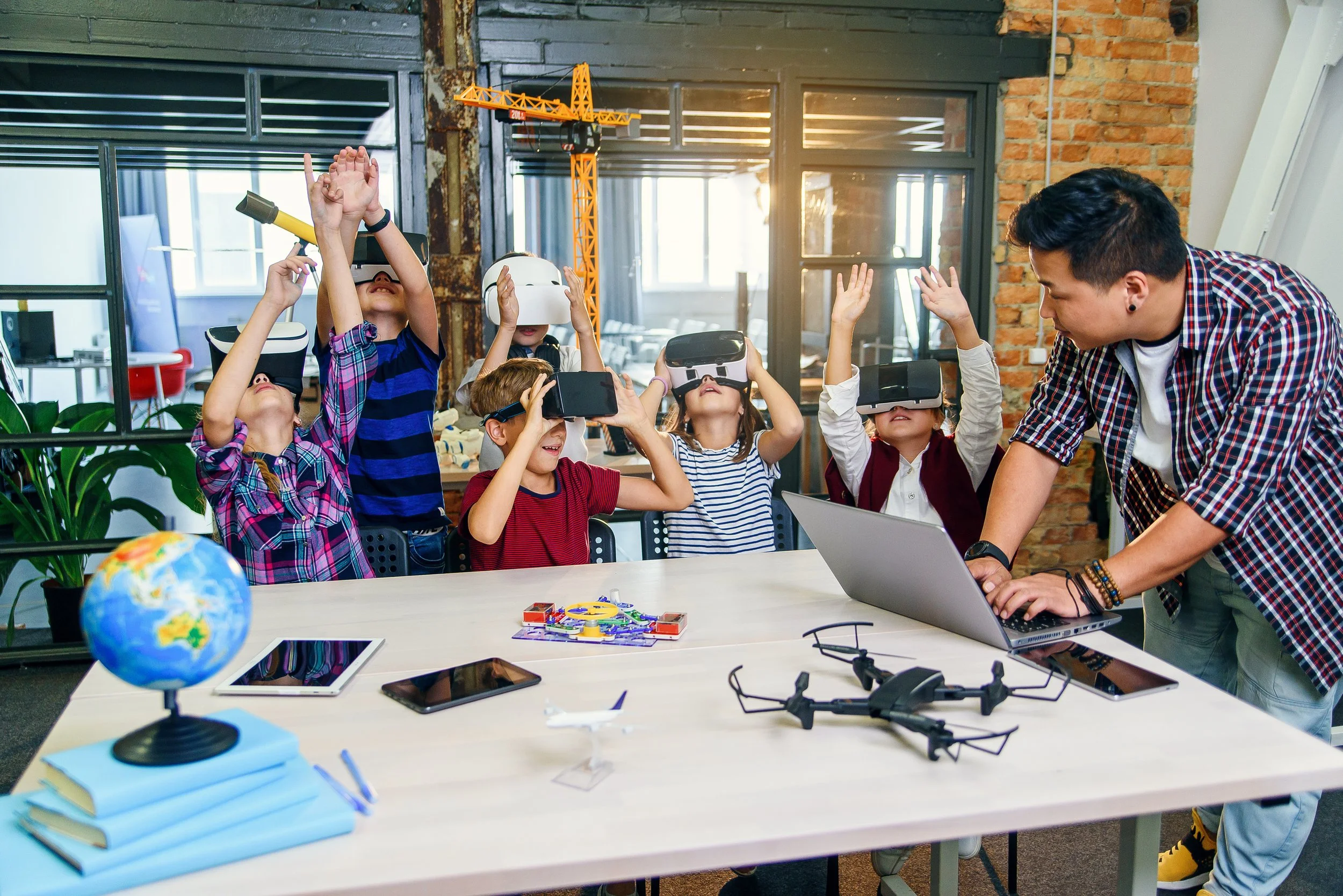 Group of children wearing virtual reality headsets engaging with a man at a table inside a modern classroom or workshop space. The table has notebooks, tablets, a drone, and a globe. The room features brick walls, large windows, and a construction cr