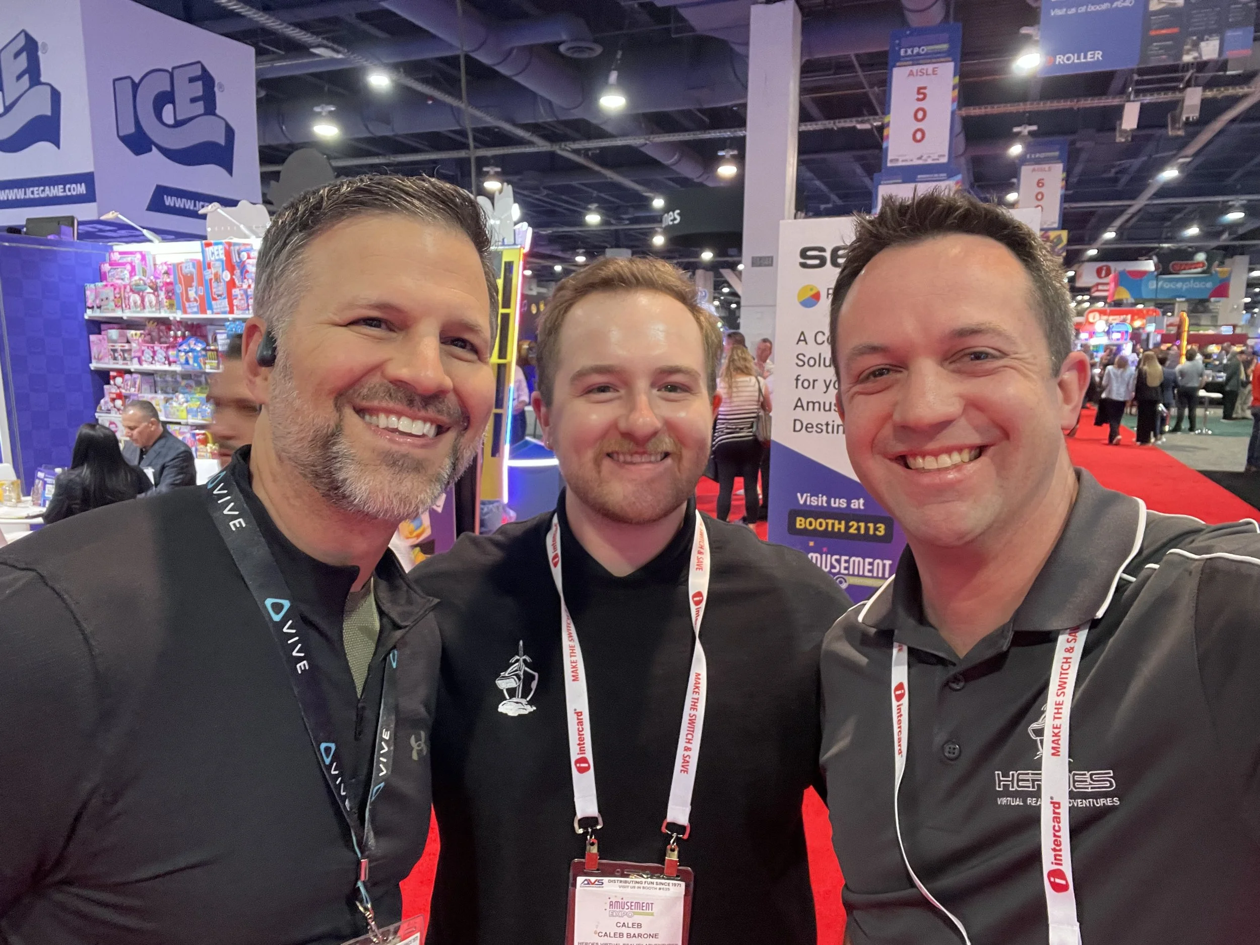 Three smiling men taking a selfie at an indoor convention or expo, with colorful booths and crowd in the background.