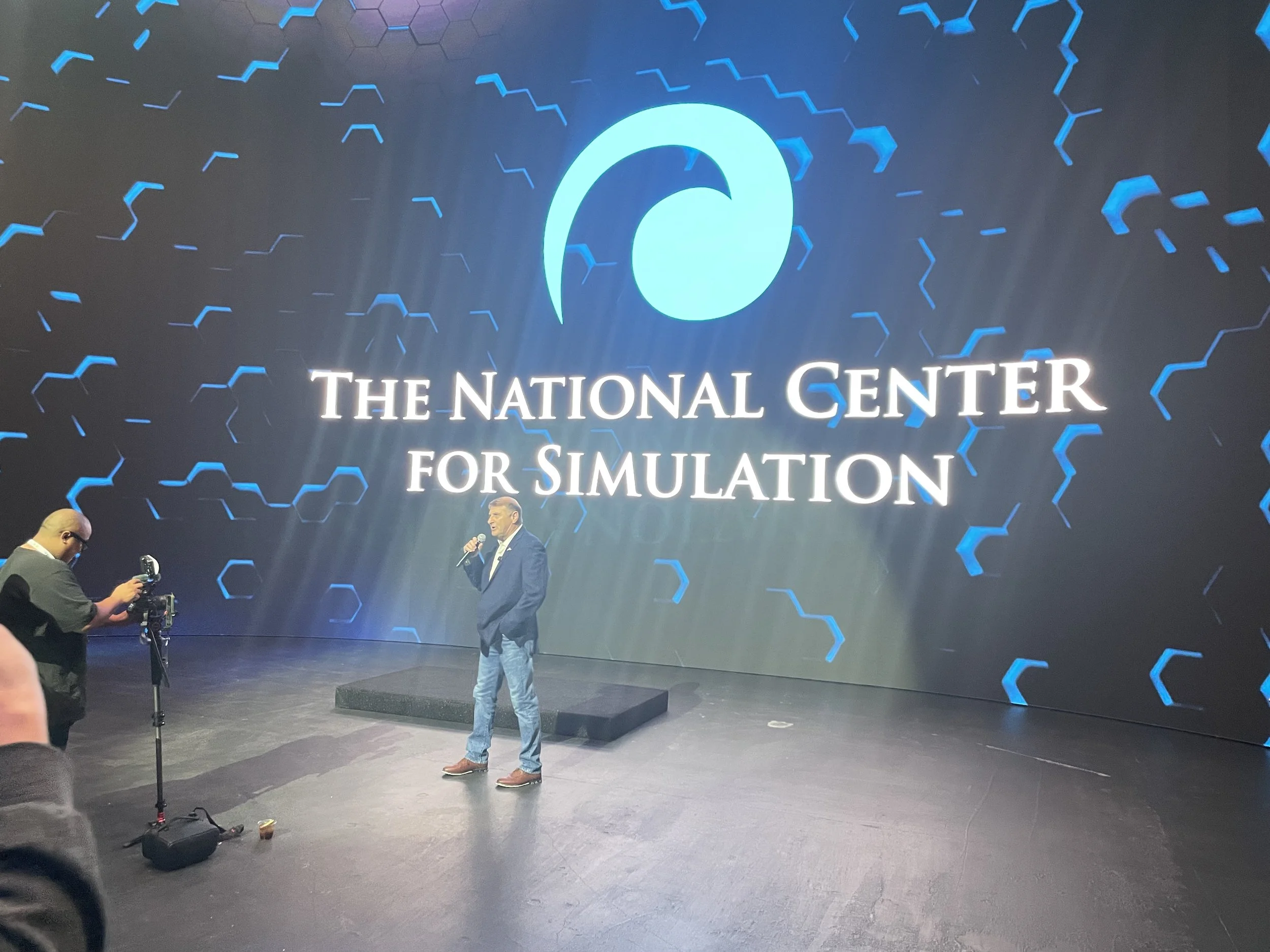 A man in a suit and jeans speaking into a microphone on stage at The National Center for Simulation event. A large screen behind him displays the organization’s logo and name, with a person taking photos on the left.