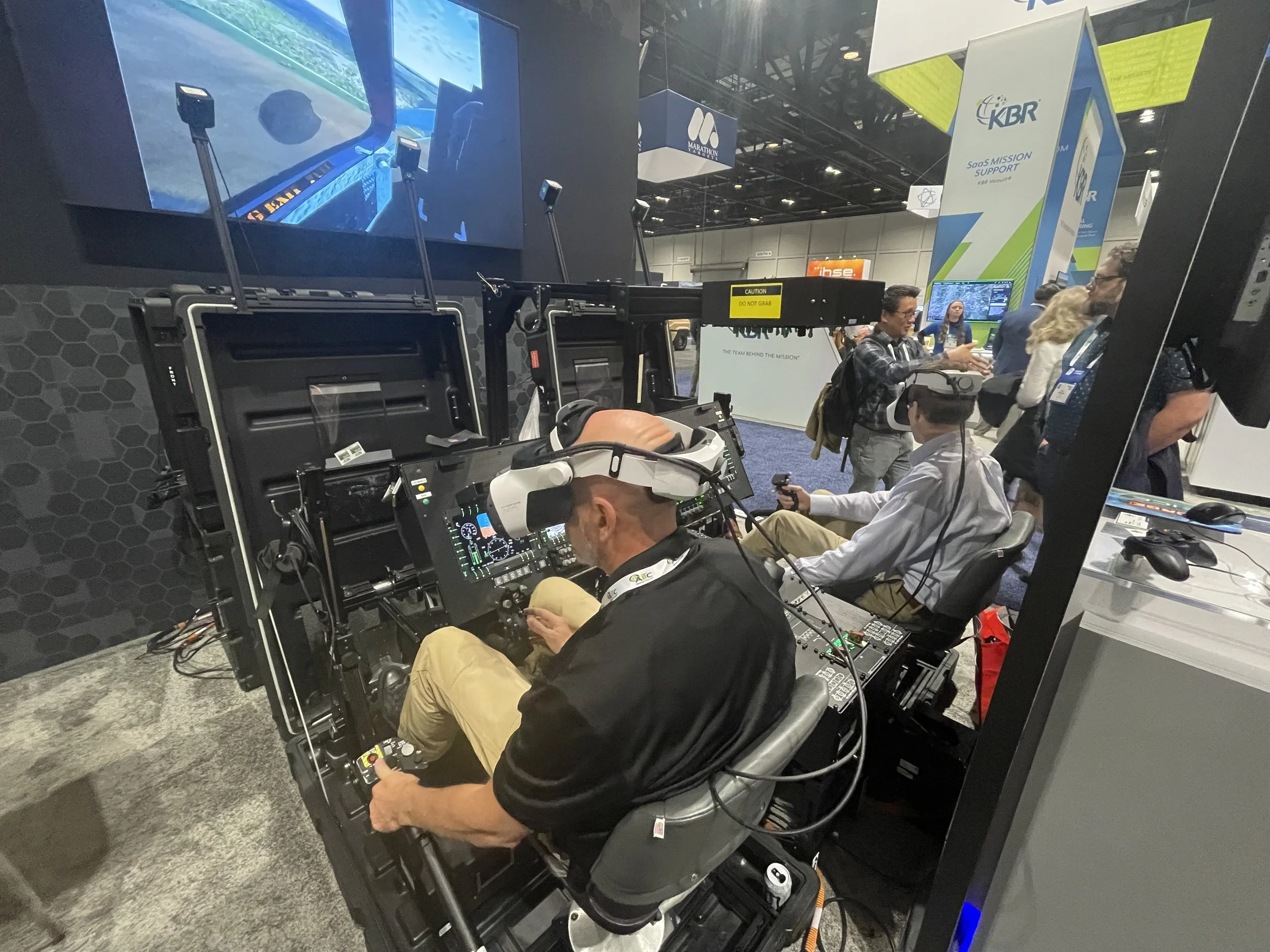 A man wearing a virtual reality (VR) headset is seated in a cockpit simulator at a technology exhibition. There are other people nearby, some engaging with different exhibits and displays.