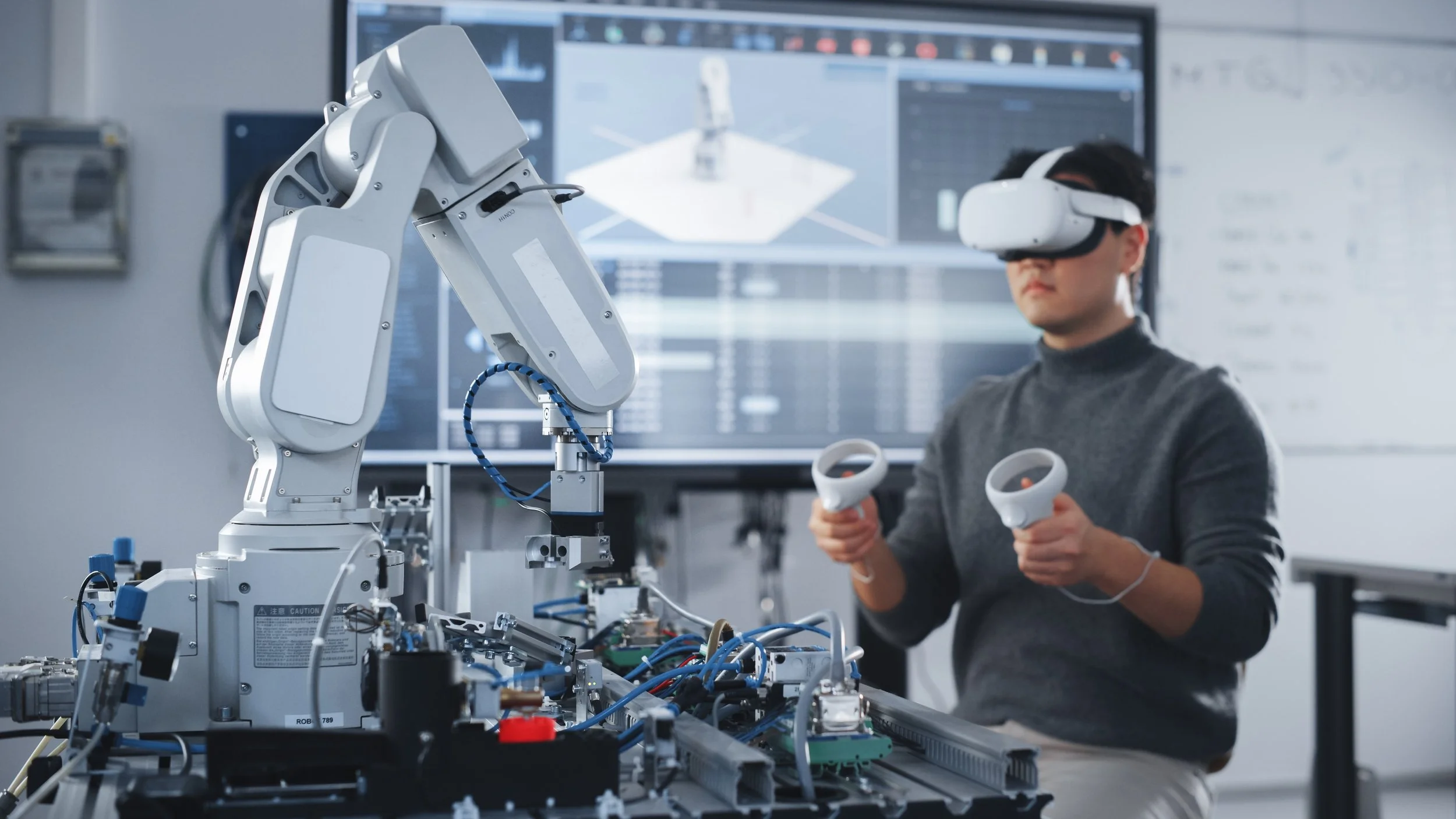 Person using a virtual reality headset and controllers while operating industrial robotic equipment in a high-tech laboratory.