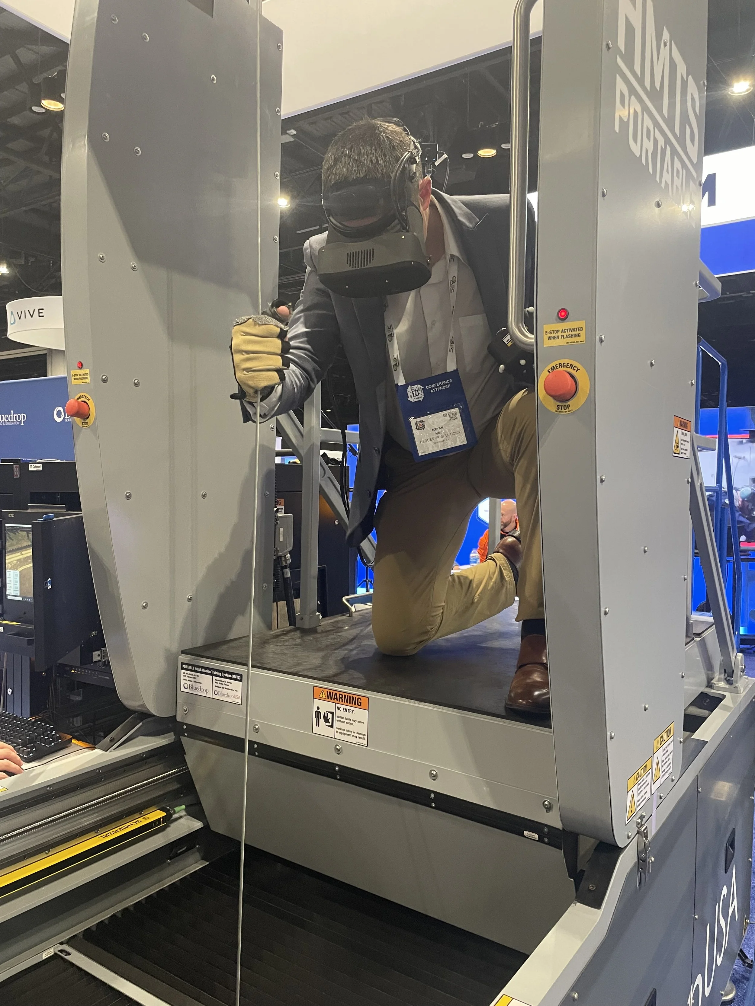 A man wearing a virtual reality headset and gloves is kneeling inside a large, enclosed virtual reality flight simulator pod. He appears to be engaging with a virtual environment.