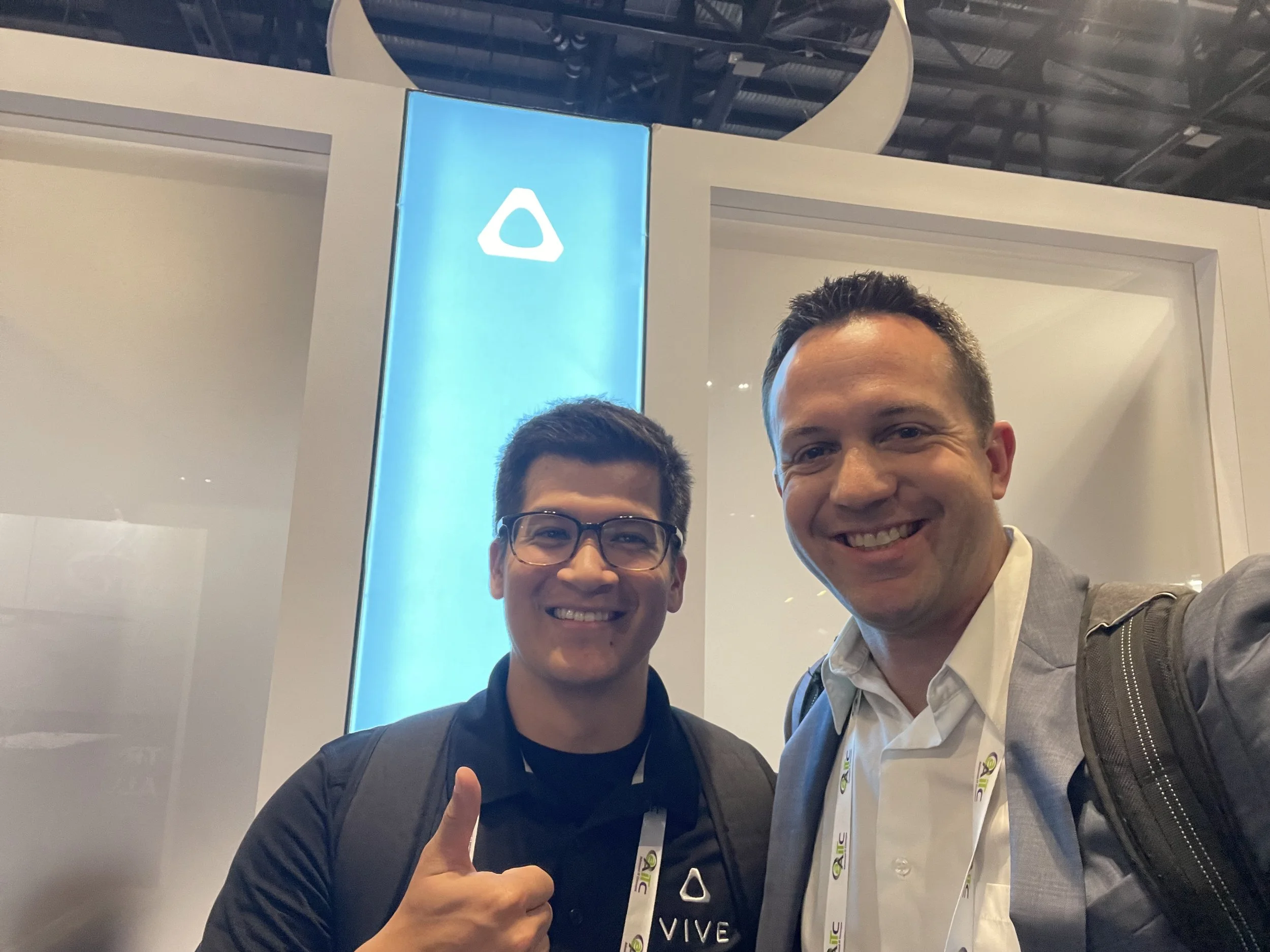 Two men smiling, one giving a thumbs up, standing in front of a blue vertical display with a white logo, at a conference or event.