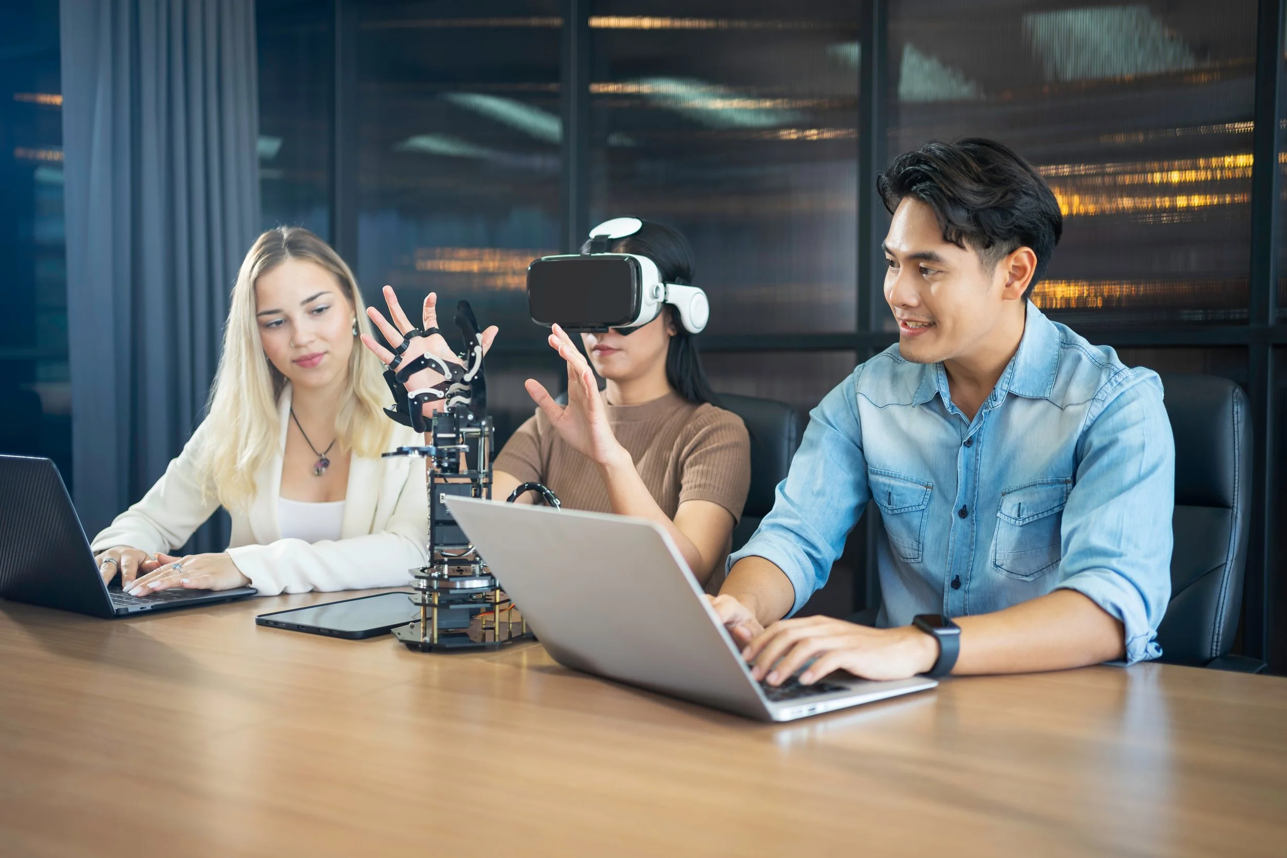 Three people working on a robotics project at a conference table in a modern office, with a robot arm, laptops, and a VR headset.