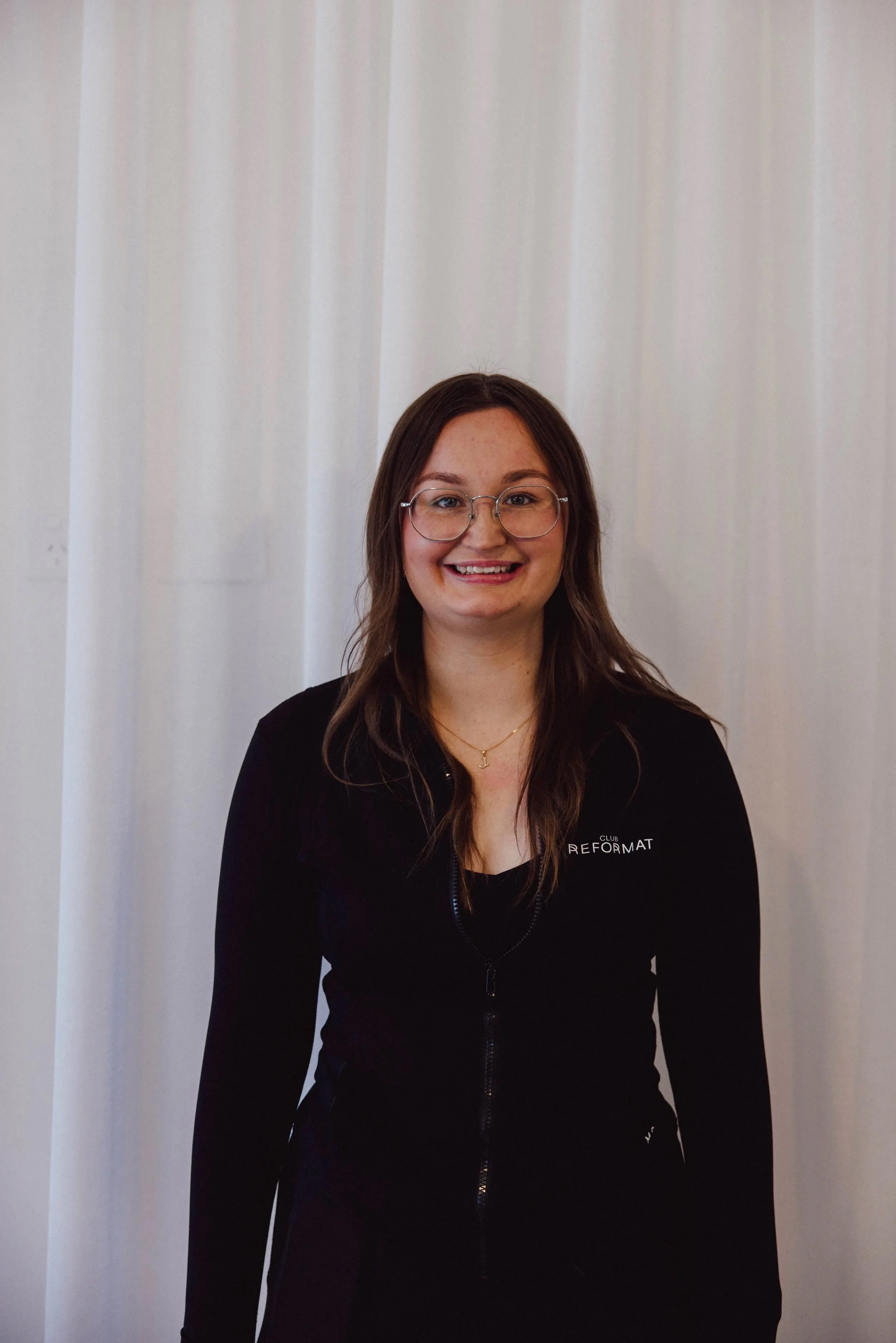 A young woman with long, wavy brown hair and glasses smiling at the camera, wearing a black zip-up jacket and gold jewelry against a plain white background.