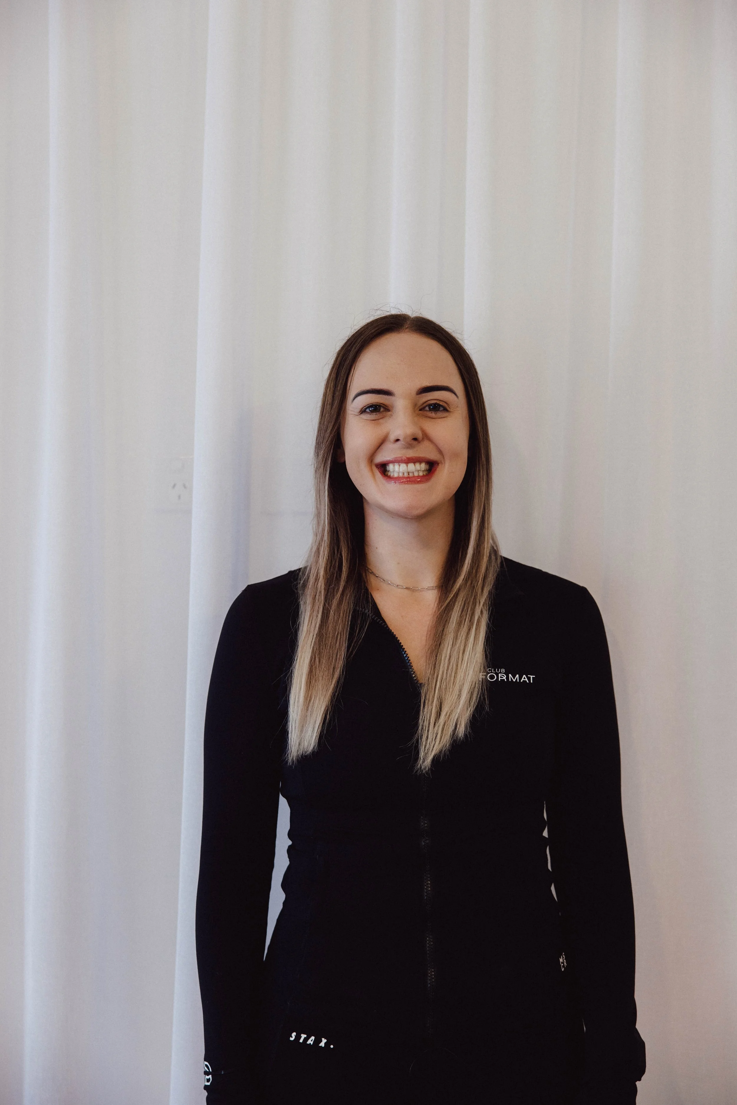Smiling woman with shoulder-length black hair wearing a blue top, standing in a lush indoor greenhouse with green plants in the background.