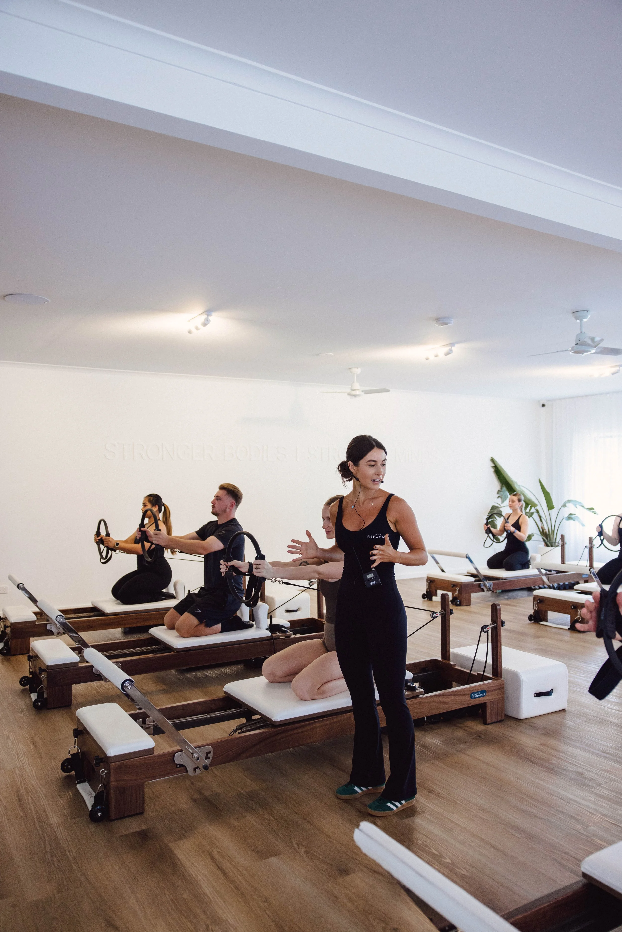 People participating in a Pilates class, lying on their backs with legs raised and arms extended, in a room with wooden wall paneling and a window.