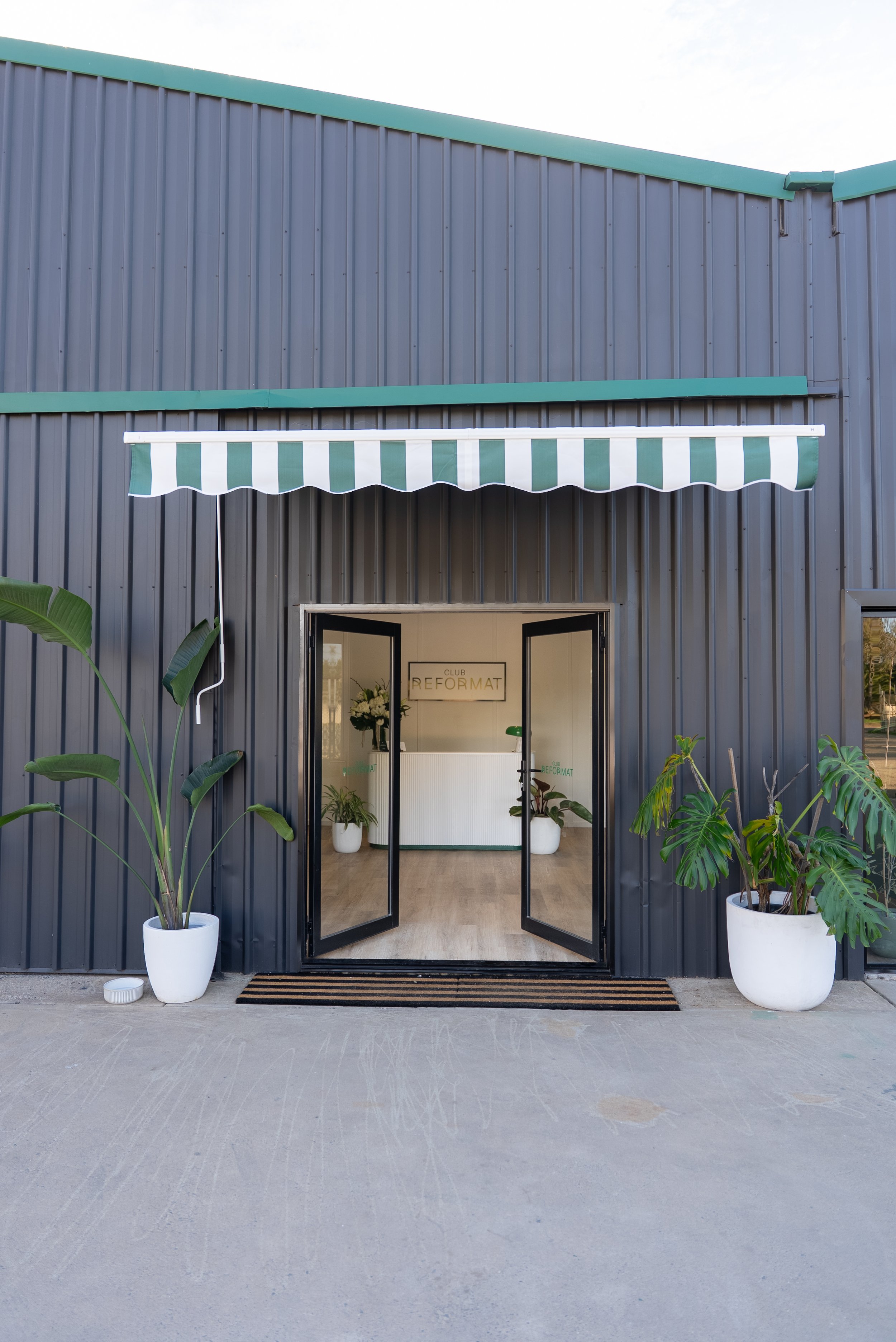The entrance to a modern building with a black framed glass double door, flanked by large potted plants, with a striped green and white awning above, and a reception desk visible inside with a sign that reads 'CLUB REFORMAT'.