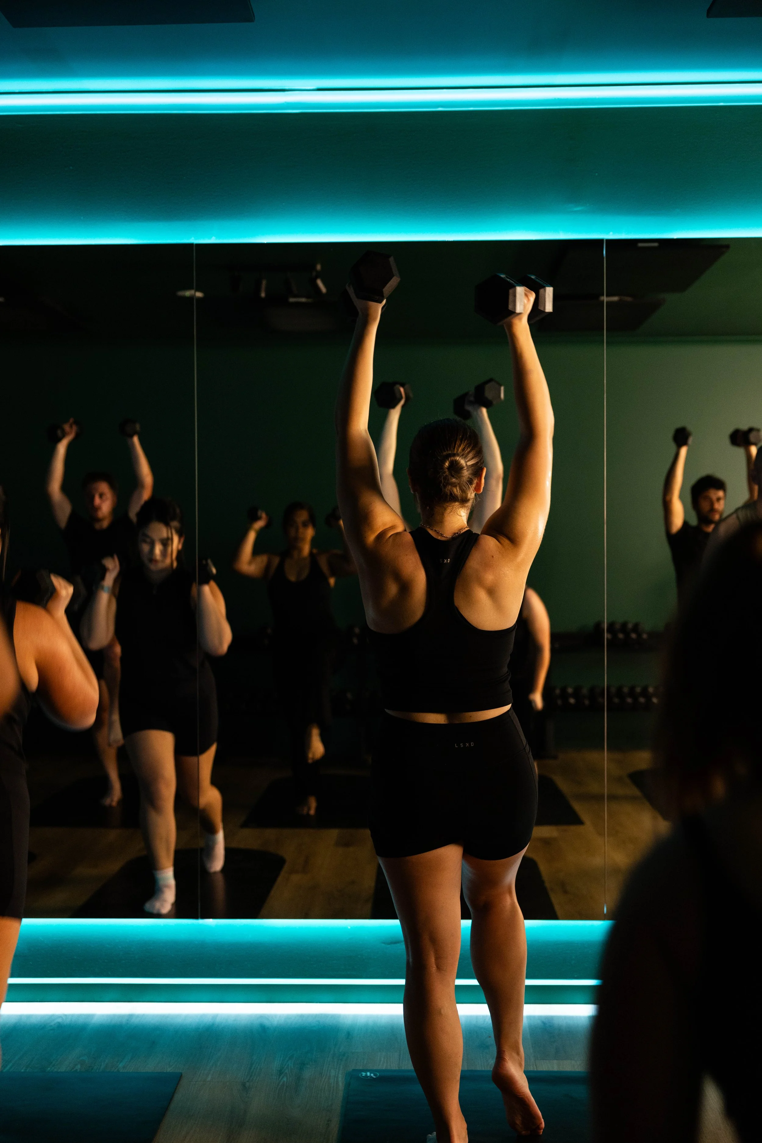 People working out in a fitness pilates class, lifting dumbbells in front of a mirror with blue lighting.