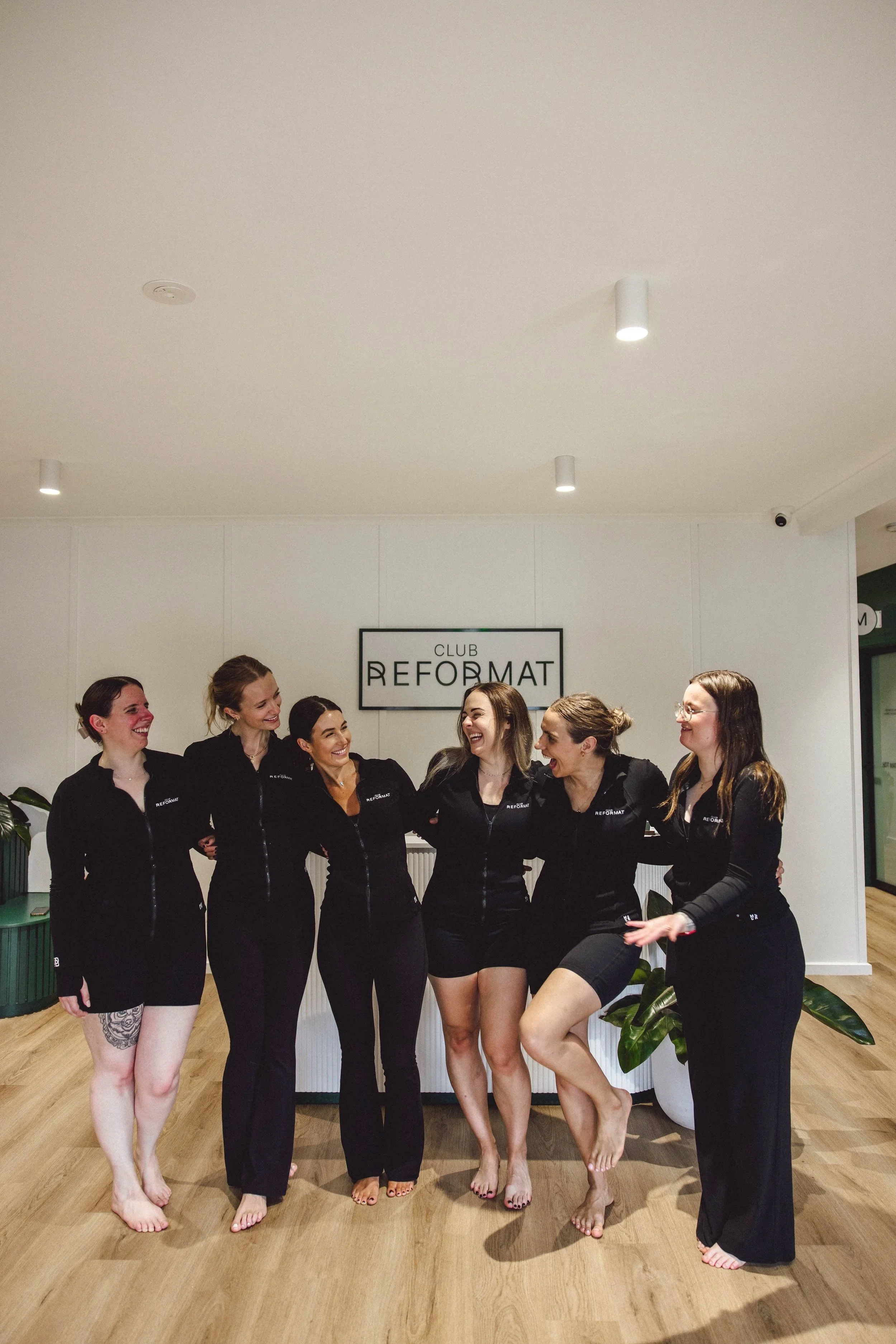 Six pilates instructors in black uniforms smiling and laughing together indoors, standing on a wooden floor with plants and a white wall behind them, and a sign that says 'CLUB REFORMAT' on the wall.
