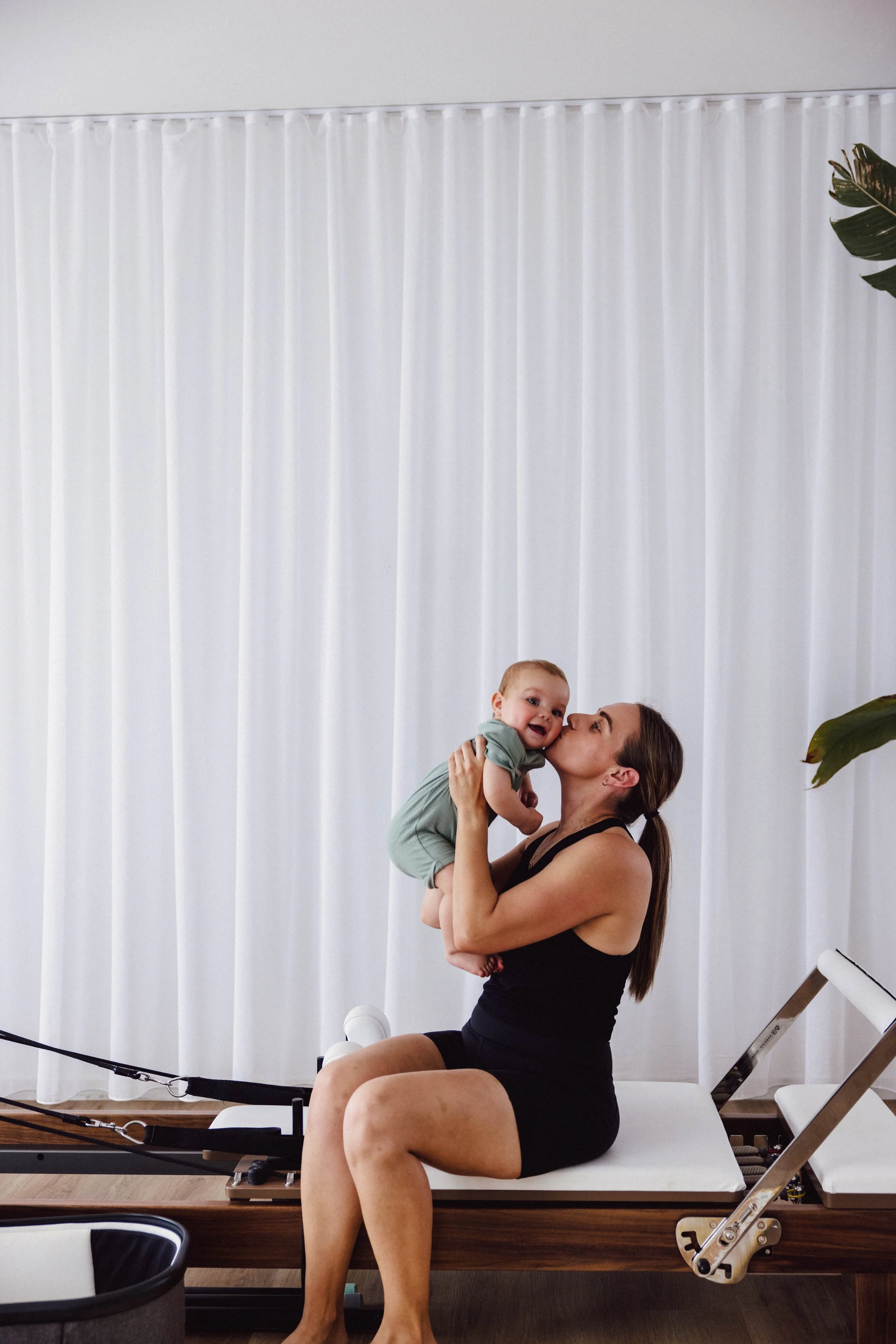 A woman in black workout clothes sitting on a reformer Pilates machine, holding a smiling baby dressed in green, in front of white curtains at a fitness studio.