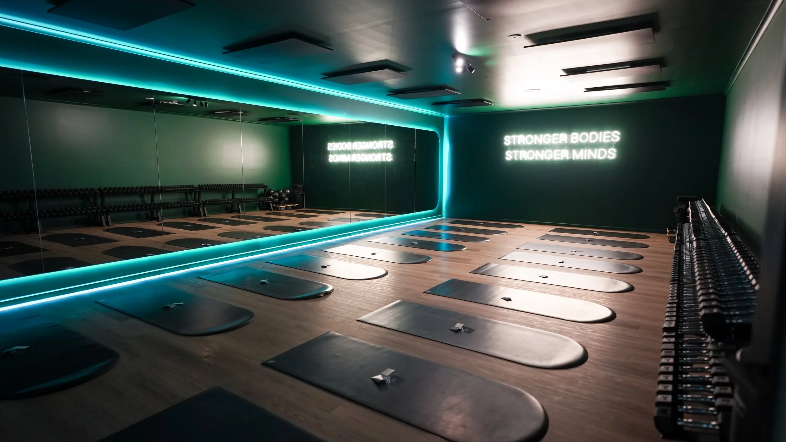 Empty hot mat pilates room with black exercise mats on wooden floor, dumbbells on the right, and a mirrored wall reflecting the room. Neon sign on the wall reads 'Stronger Bodies, Stronger Minds'.