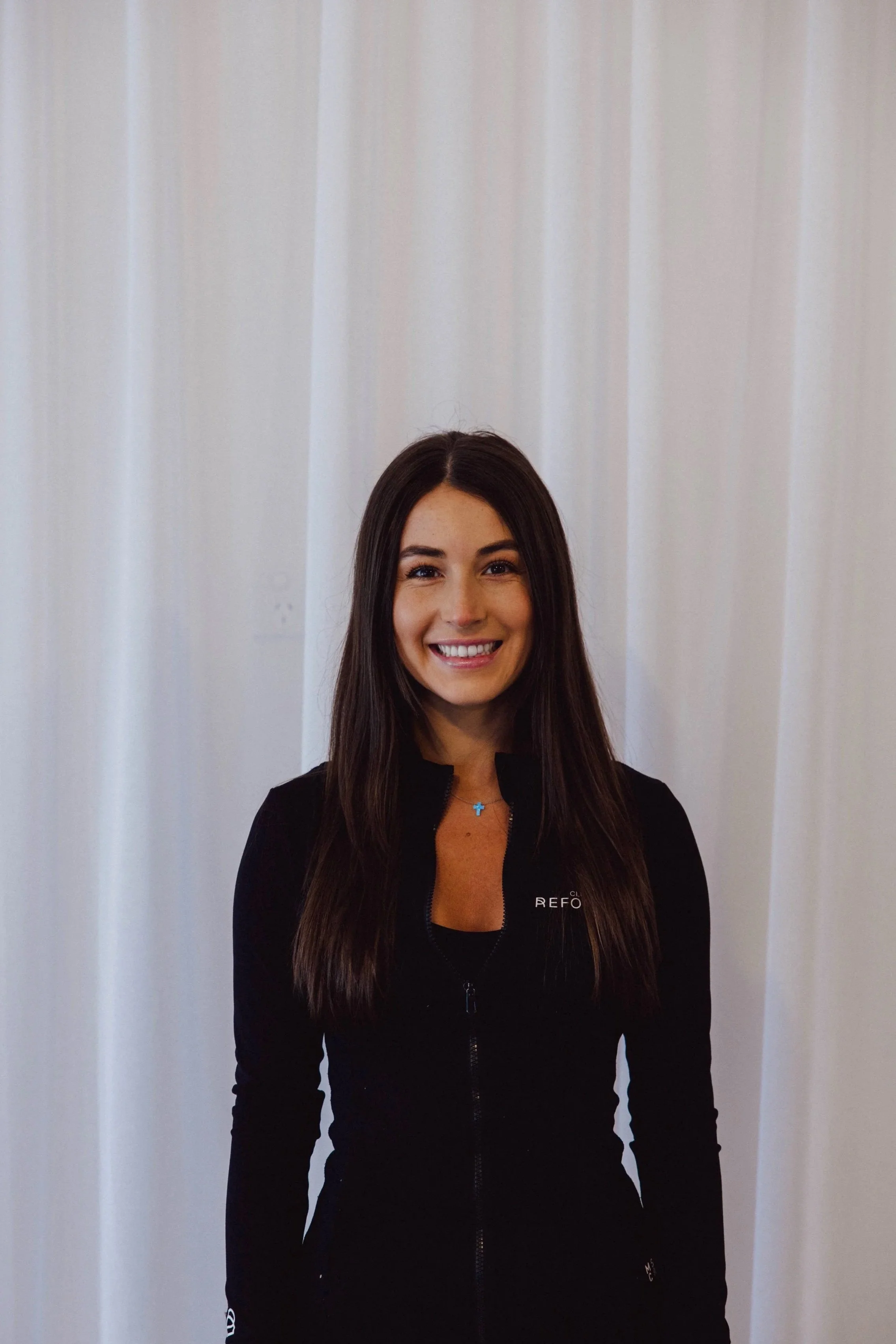 A young woman with long brown hair and a bright smile, wearing a black zip-up top, standing against a white background.