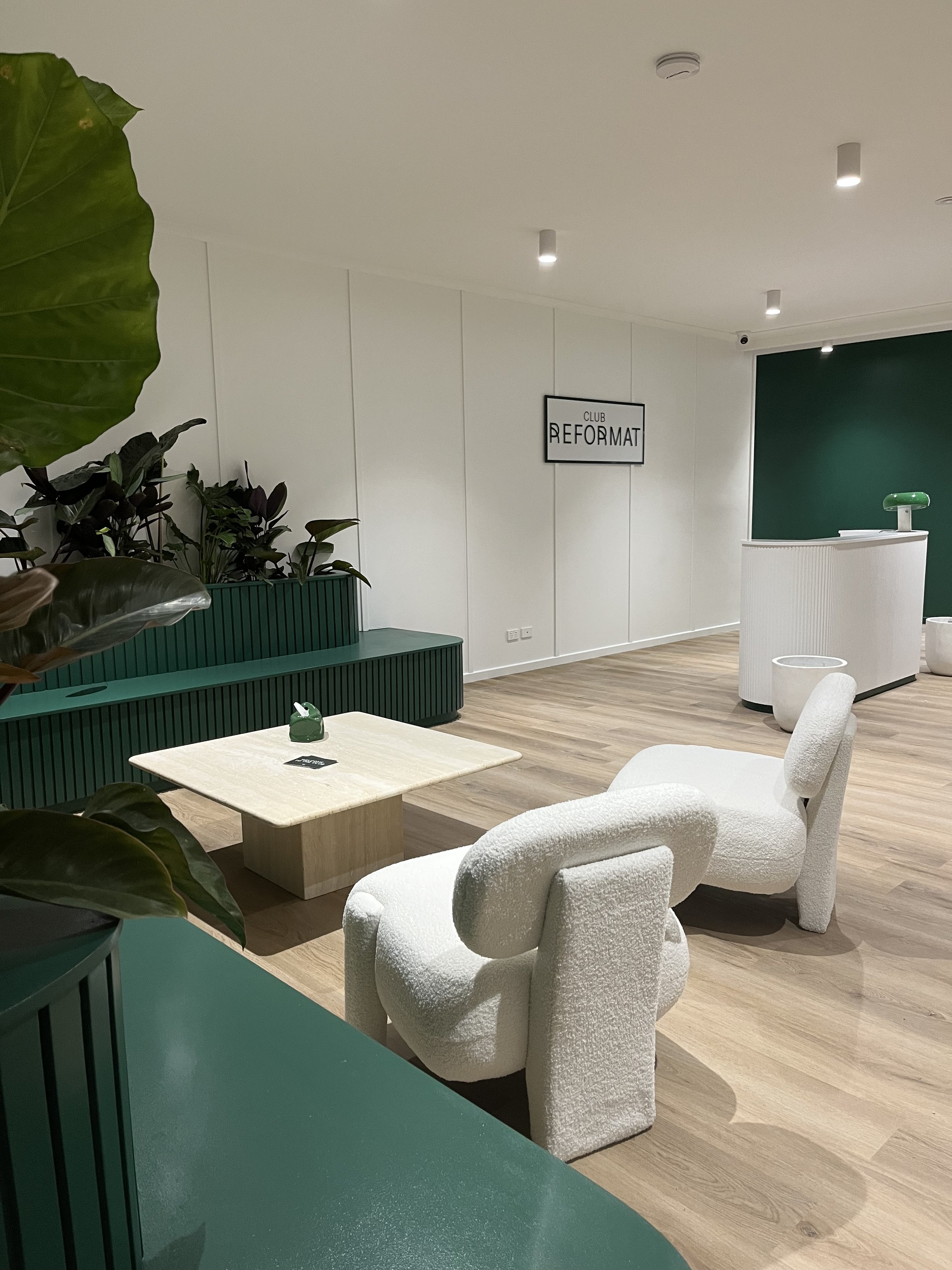 Modern hotel or club reception area with green and white interior, featuring white textured armchairs, a small light-colored coffee table, green cabinetry, and a large green wall with a black and white sign reading "Club Reformat". There are decorative plants and a curved reception desk in the background.