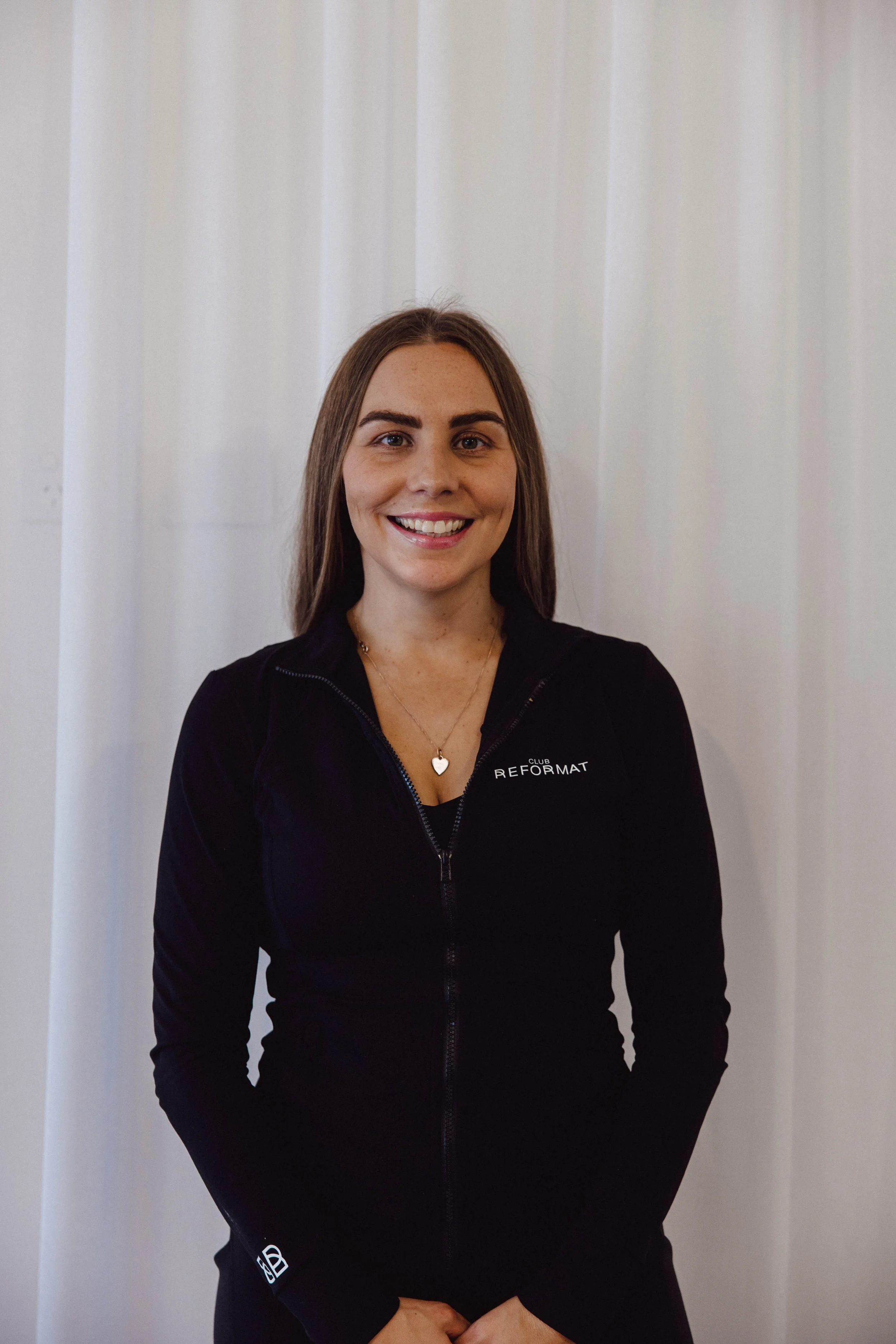 Close-up of a man with curly dark hair, glasses, and a beard, wearing a dark green sweater, looking to the right with a slight smile, in a room with a white wall and a green plant in the background.