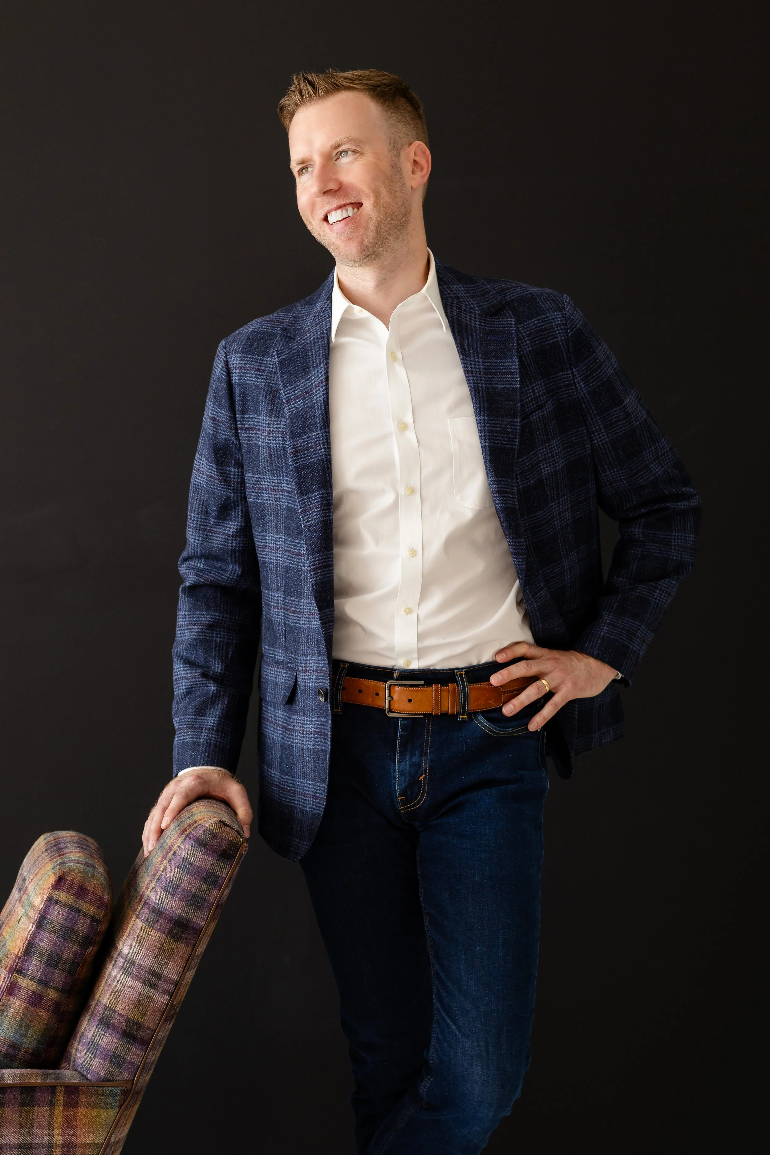 A man with short hair and a beard smiling, dressed in a white shirt, blue plaid blazer, dark jeans, and a brown belt, standing next to a patterned chair against a dark background.