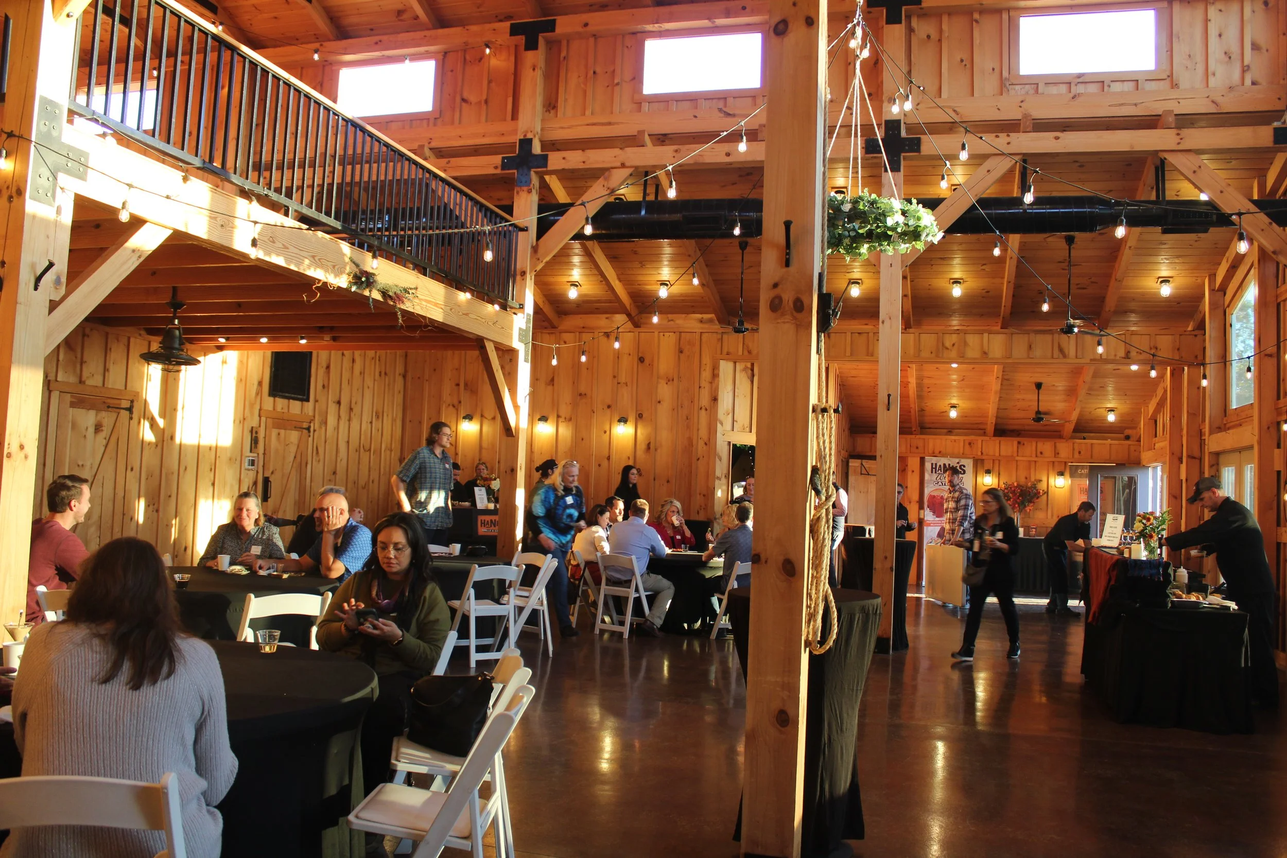 People sitting and standing in a rustic wooden indoor event space with string lights, high windows, and wooden beams.