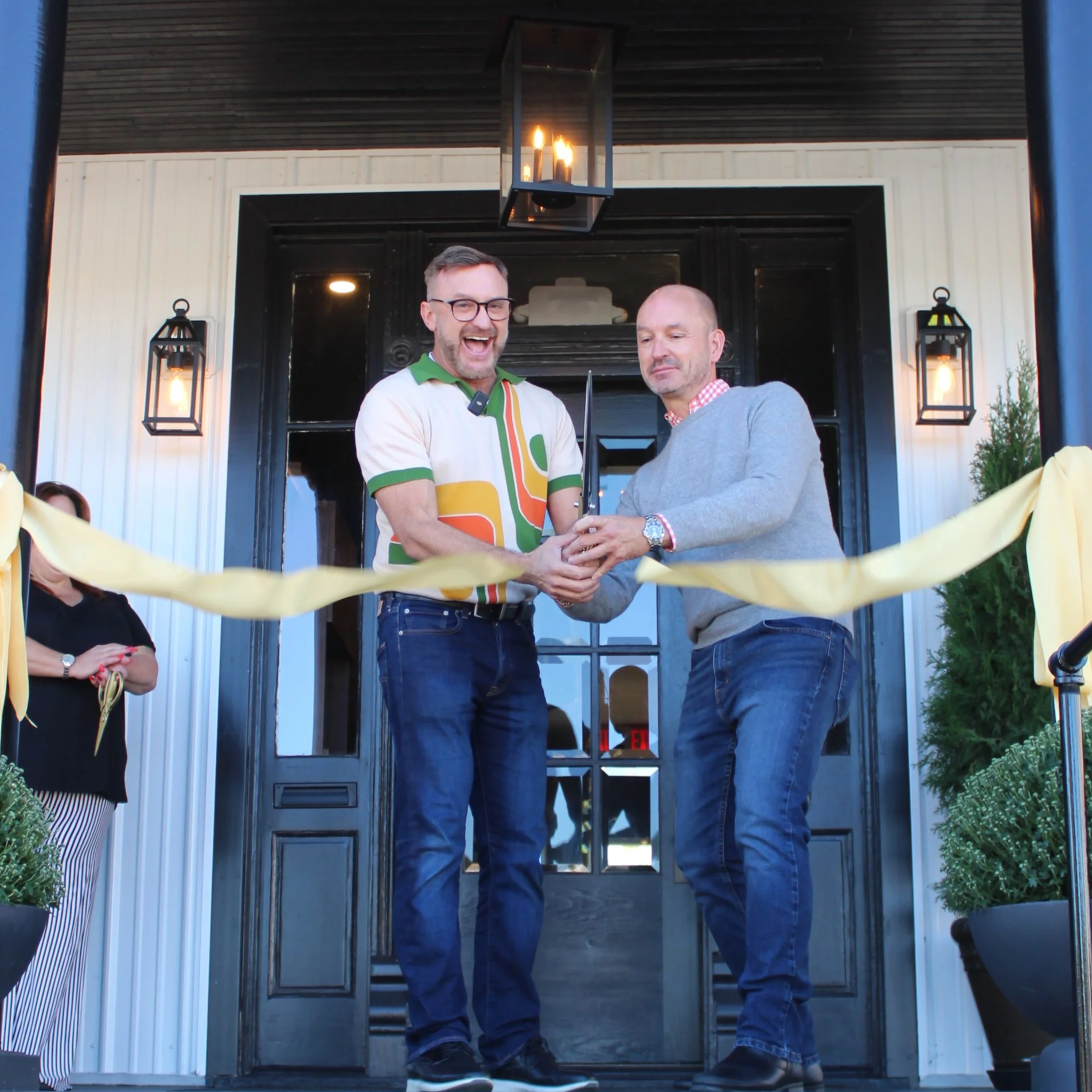 Two men stand in front of a black front door with glass panels, holding a ceremonial knife together during a celebration or special occasion outside a house, with a yellow ribbon stretched across the entrance.