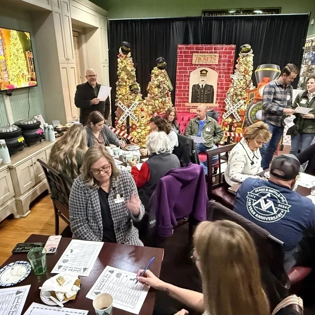 People dining in a restaurant with a railway theme, decorated with a backdrop of vintage railroad signs, trees made of flowers, and a large model train in the background.