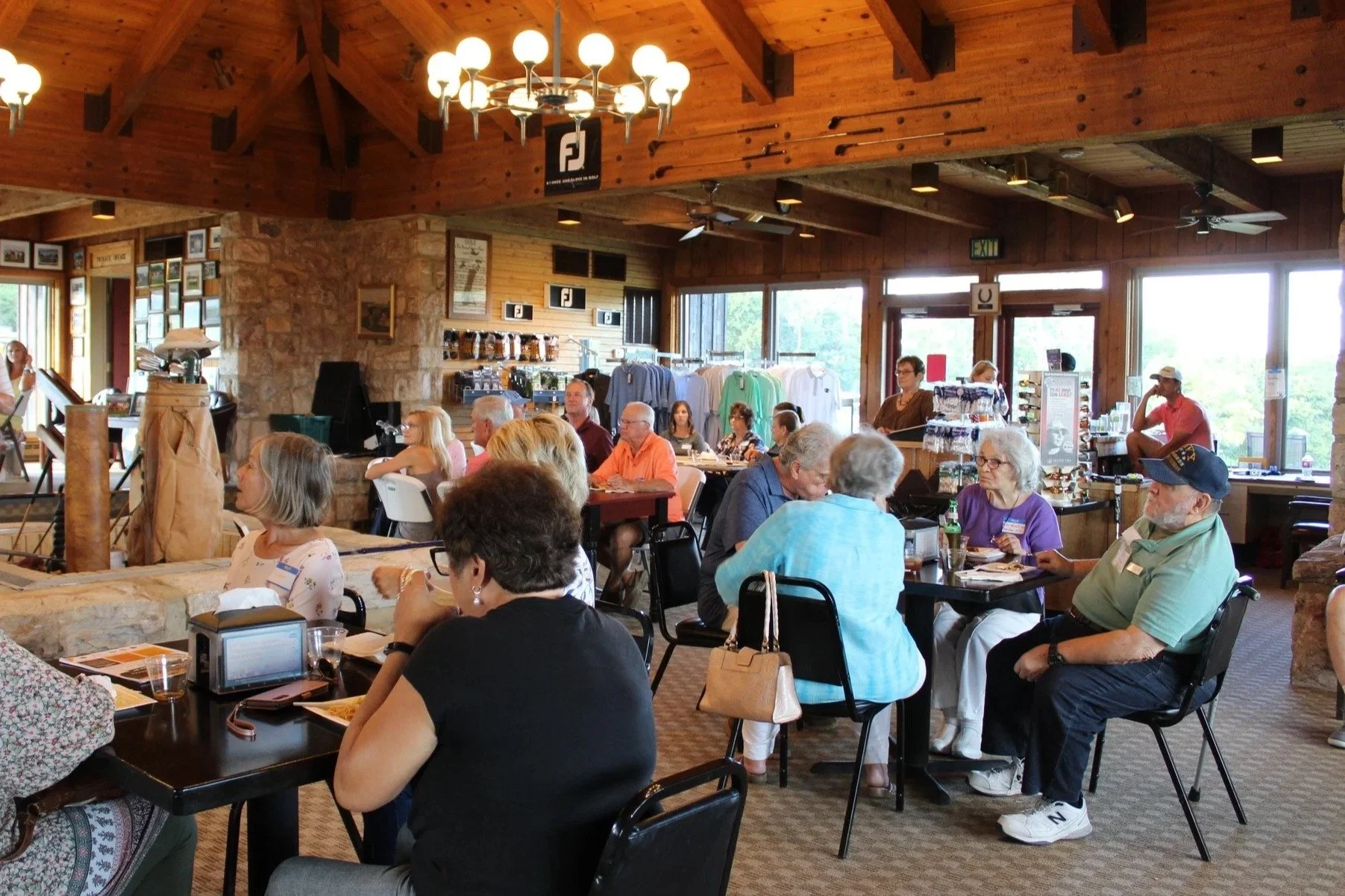 A group of people gathered inside a rustic, wood-paneled restaurant or cafe, seated at tables, some eating and others talking. The room has large windows letting in natural light, and there are various clothing and merchandise displays in the backgro