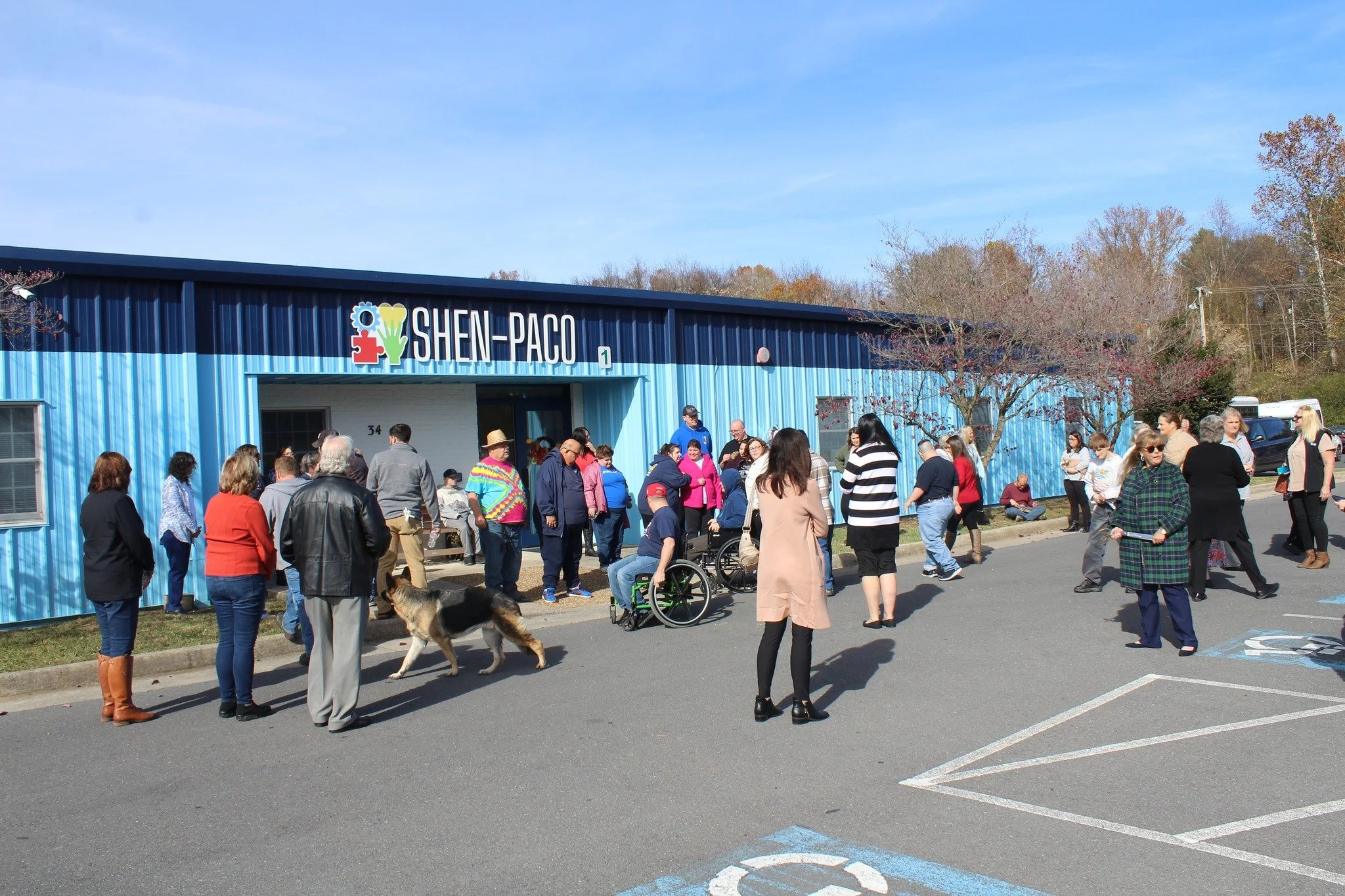 Group of people gathered outside a blue building with a sign that reads 'SHEN-PACO,' some in wheelchairs, standing and walking on a sunny day with trees in the background.