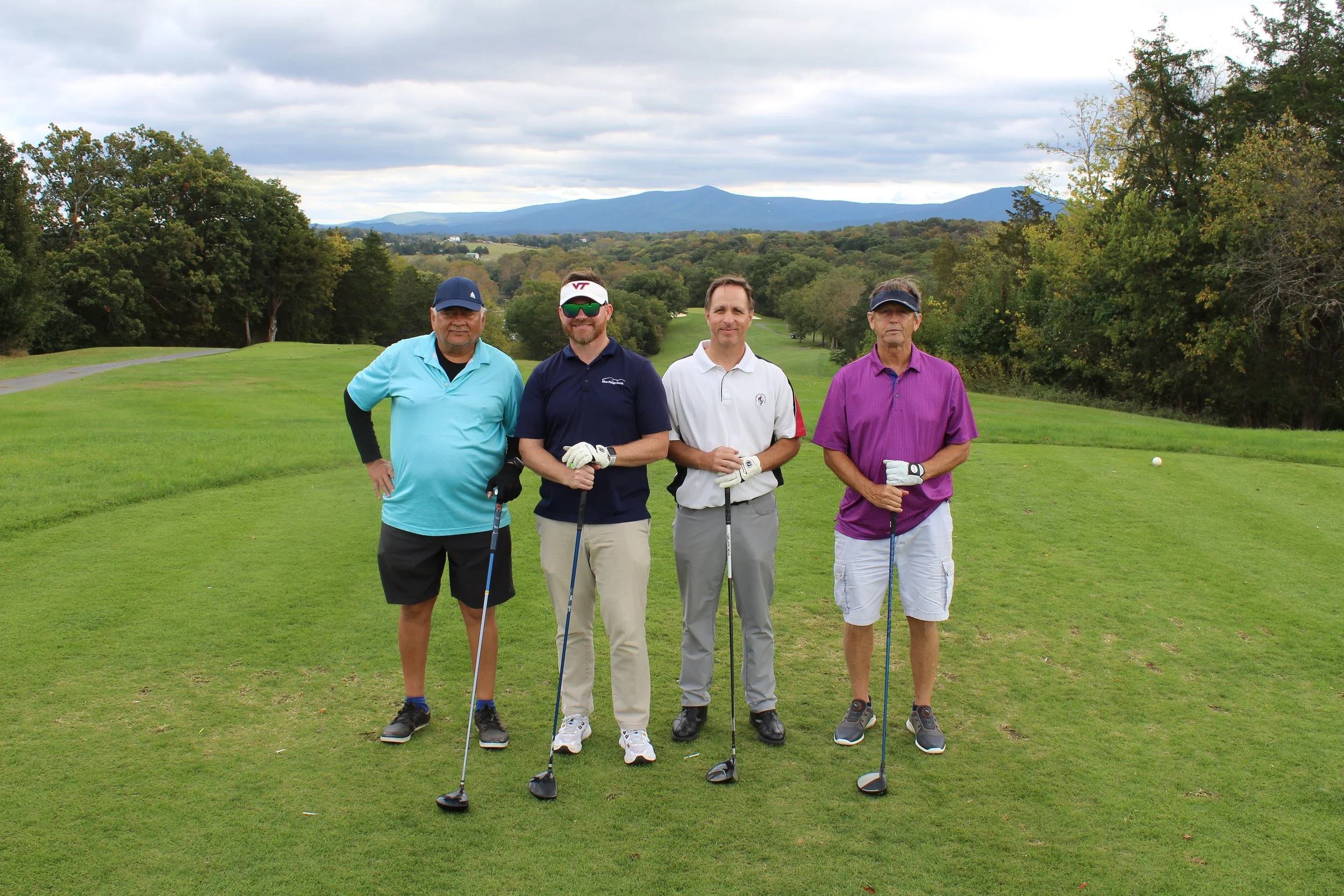 Four men standing on a golf course with trees and mountains in the background, holding golf clubs.