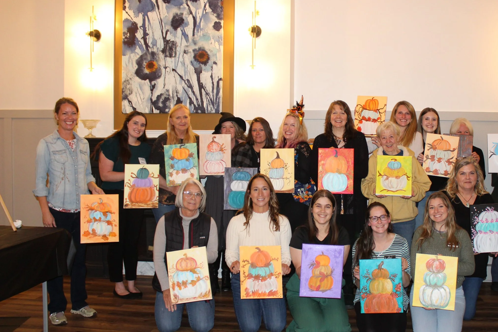 Group of women posing with pumpkin-themed paintings at an art class or event.