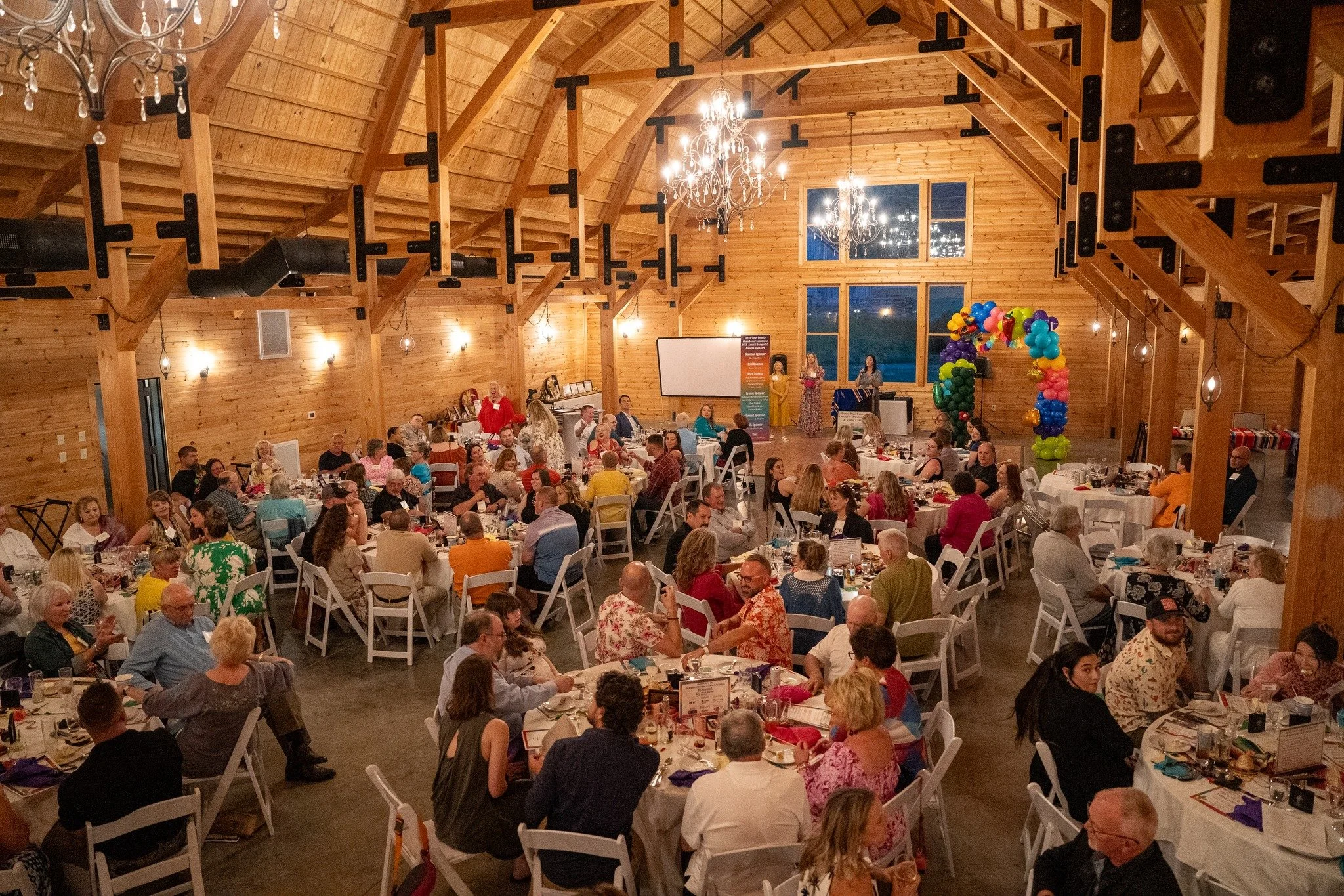 A large wooden banquet hall decorated for an event, with many tables filled with people, a large window, hanging chandeliers, and colorful balloon decorations.